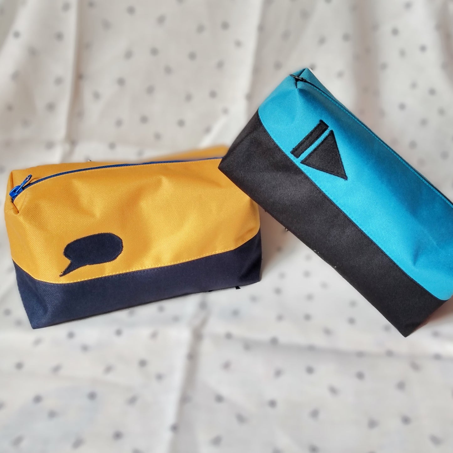 Deux trousses de toilette Corail qui se chevauchent. Elles sont rectangulaires et bicolores, en horizontal. L'une est noire en bas et bleu paon en haut, avec un appliqué "suivant" noir sur le bleu. La seconde est bleu foncé et jaune moutarde avec un appliqué en forme de bulle de bande dessinée bleu sur le jaune. Elles se ferment avec une fermeture éclair.