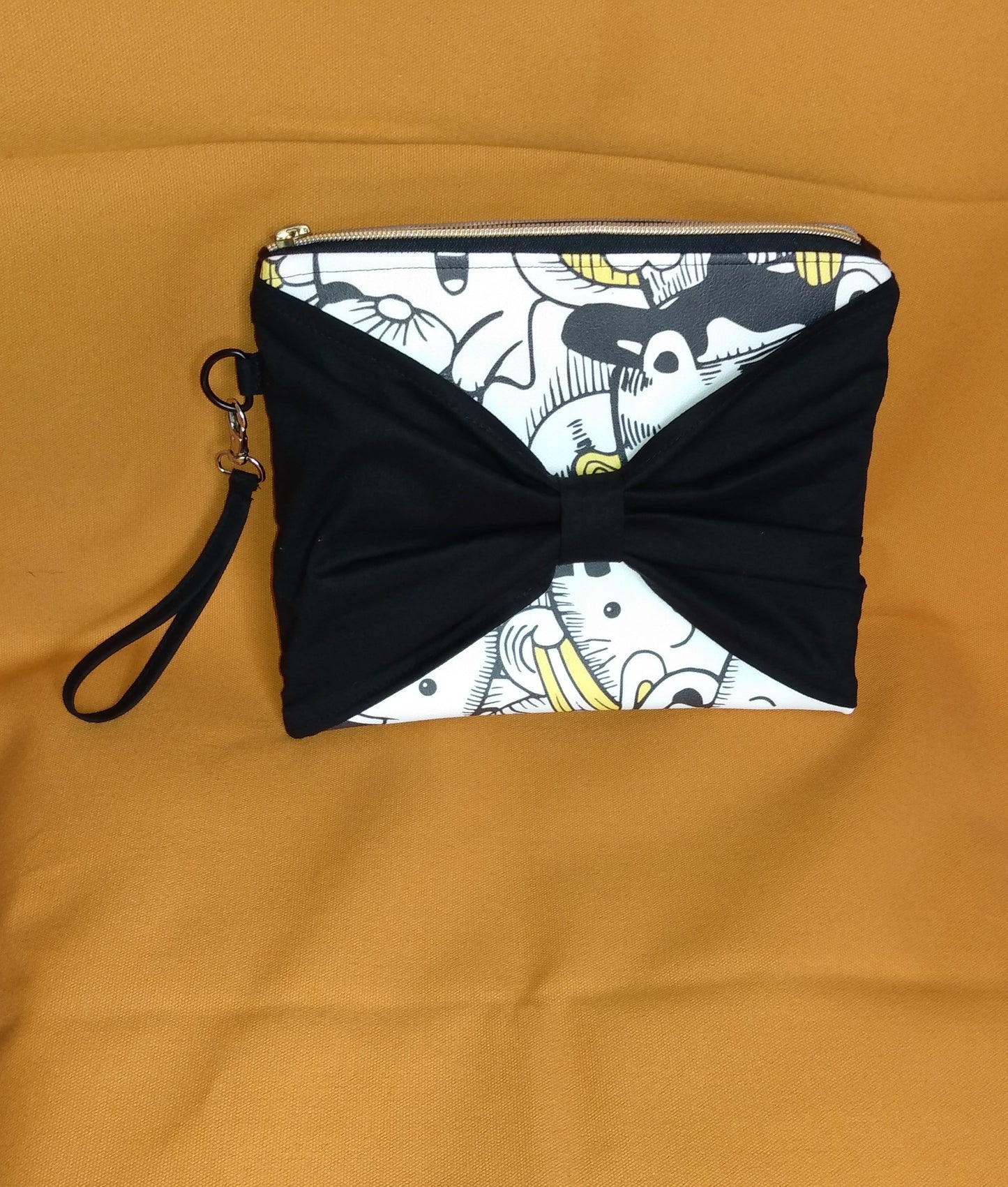 Face avant de la pochette Bonidé. Elle est en simili cuir imprimé chat blanc, noir et jaune. Un noeud est cousu sur l'avant et cache une partie de la face. Une dragonne est visible sur le côté, accrochée avec un mousqueton. La pochette se ferme en haut avec une fermeture éclair dorée.