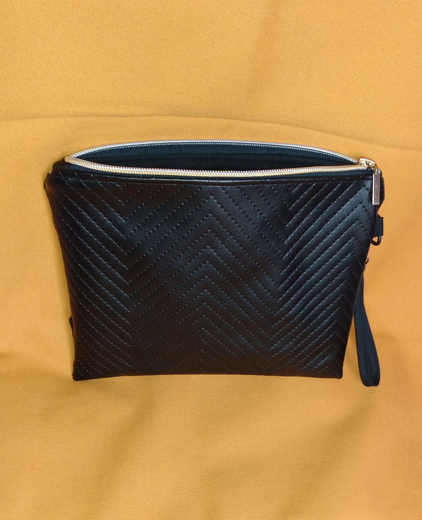 Arrière de la pochette Bonidé. Elle est en simili cuir noir à chevrons. On voit la dragonne noire sur le côté et la fermeture éclair ouverte laisse apercevoir la doublure en coton noire.