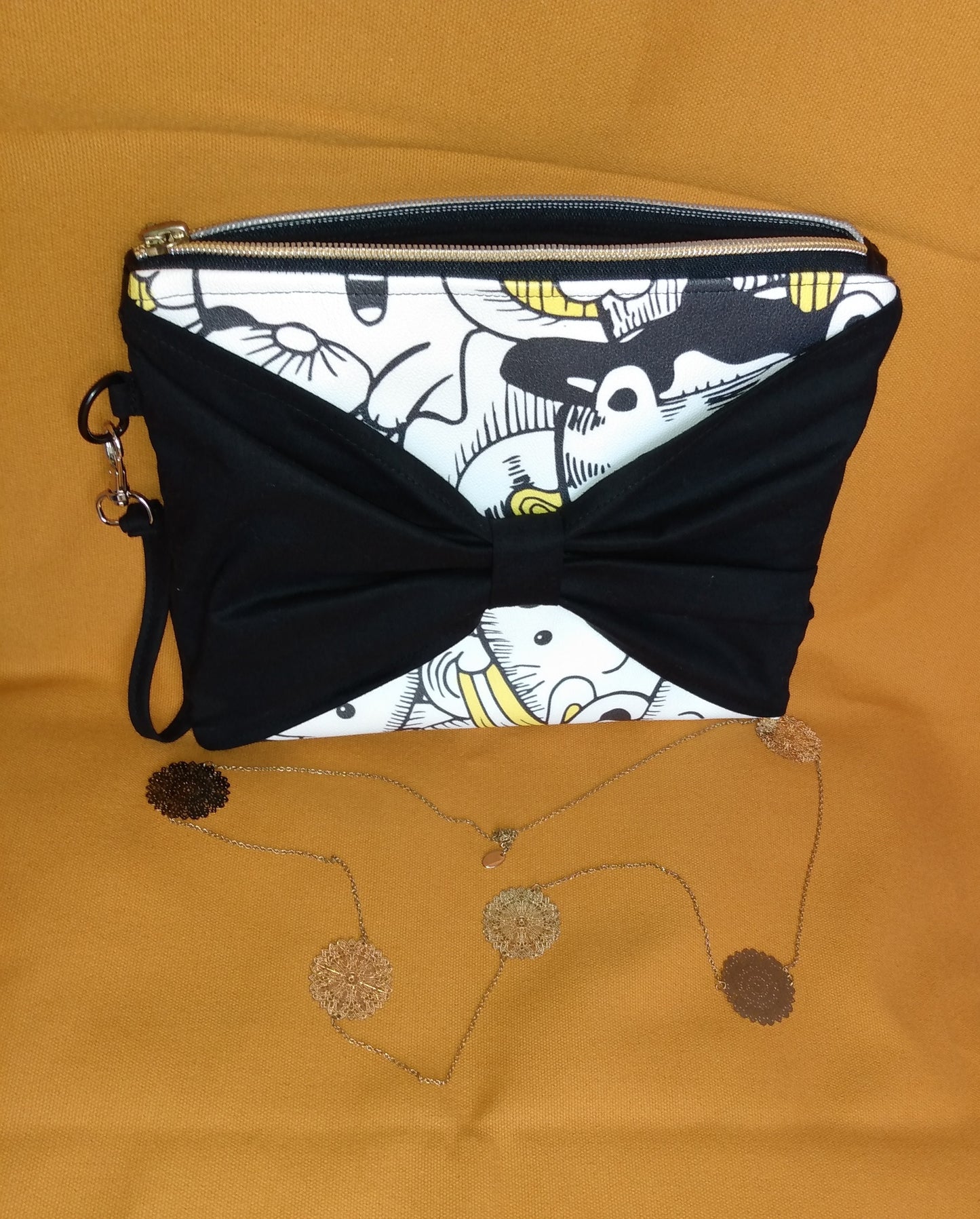 Face avant de la pochette Bonidé. Elle est en simili cuir imprimé chat blanc noir et jaune. Un noeud papillon est en avant et cache donc une partie de la face. On peut voir une dragonne noire sur le côté, accrochée par un mousqueton. La pochette se ferme avec une fermeture éclair dorée.