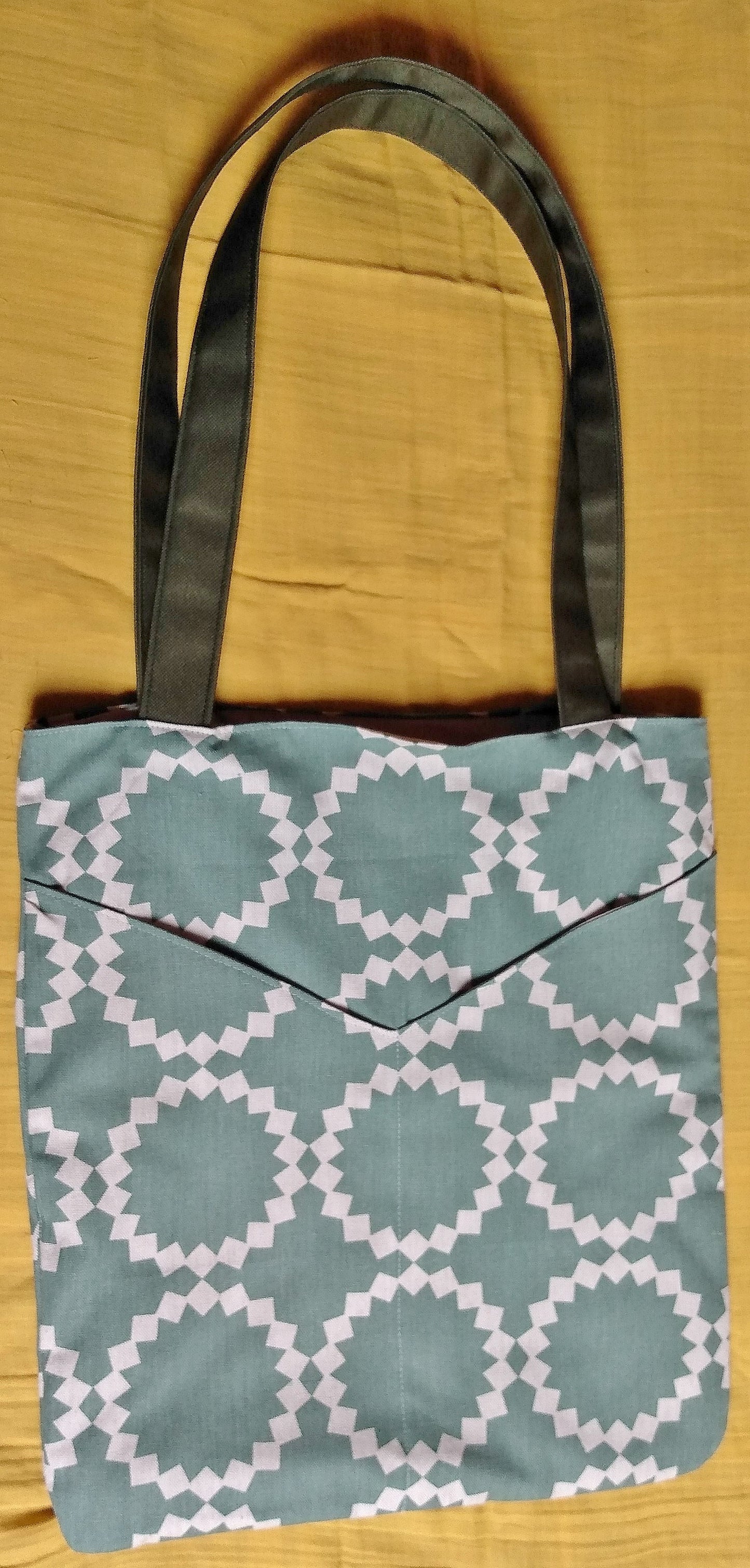 Avant du sac balad'in soleil vert. Le sac est en toile coton lin verdé avec des dessins géométriques en forme de soleil beige. On voit 2 grandes poches plaquées dans le même tissu à l'avant.
Les anses sont en toile à sac kaki.