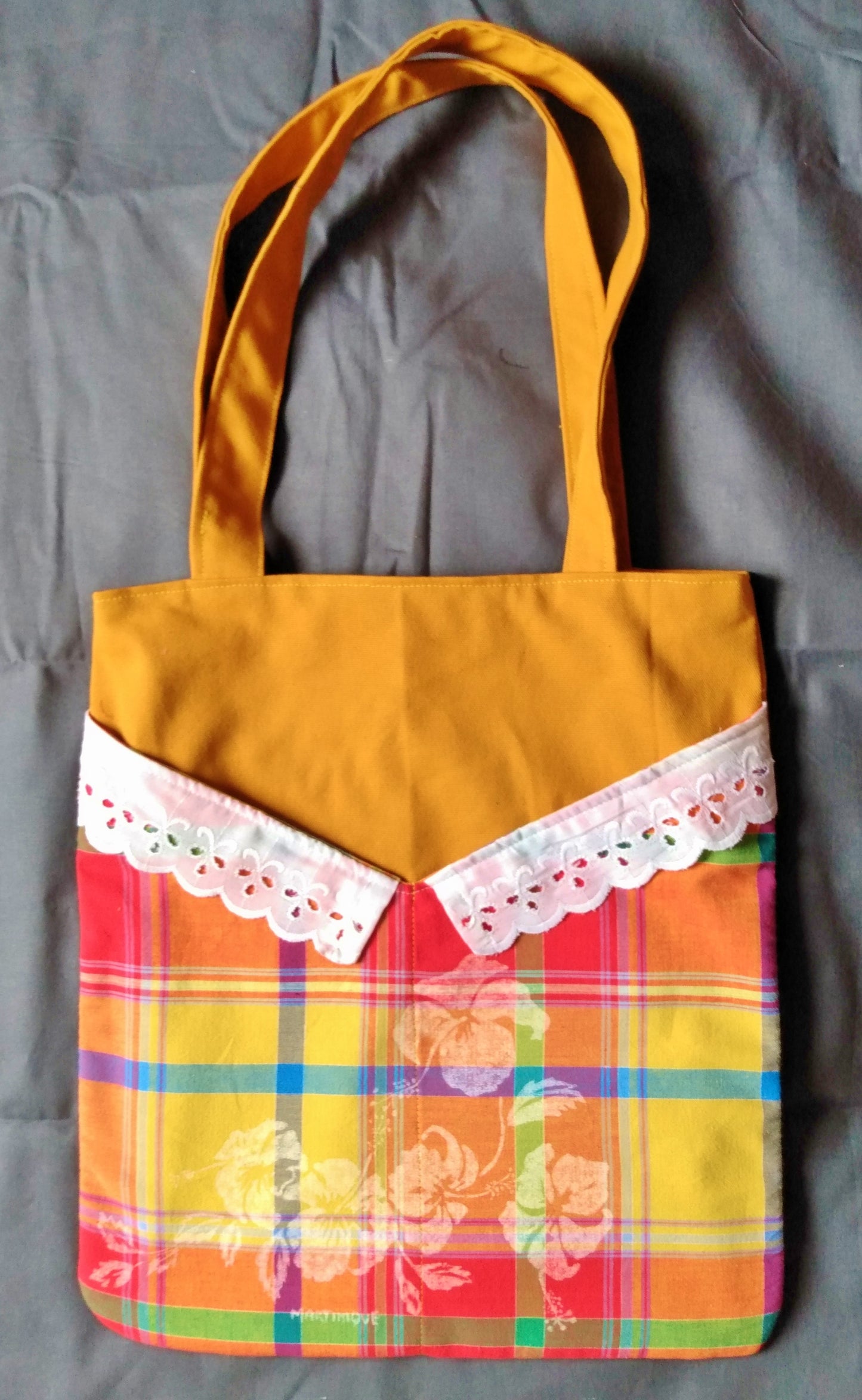 Avant du sac Balad'In, tissu principal jaune moutarde avec deux poches plaquées, qui partent du bas jusqu'aux 3/4 du sac environ. Poches motif floral à carreaux style madras, surplombé de dentelle. Les anses sont faites pour porter le sac à l'épaule et sont jaunes moutarde.