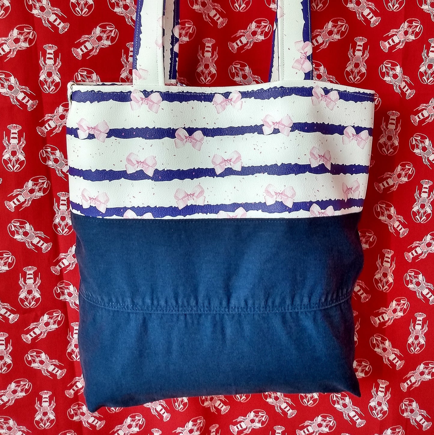 Avant du sac Jeanie. Le haut (jusquà la moitié du sac) est en simili cuir rayé blanc et bleu avec des noeuds roses. Les anses sont dans le même tissu. Le bas est constitué de deux bande de jean bleu foncé cousues ensemble à l'horizontal.