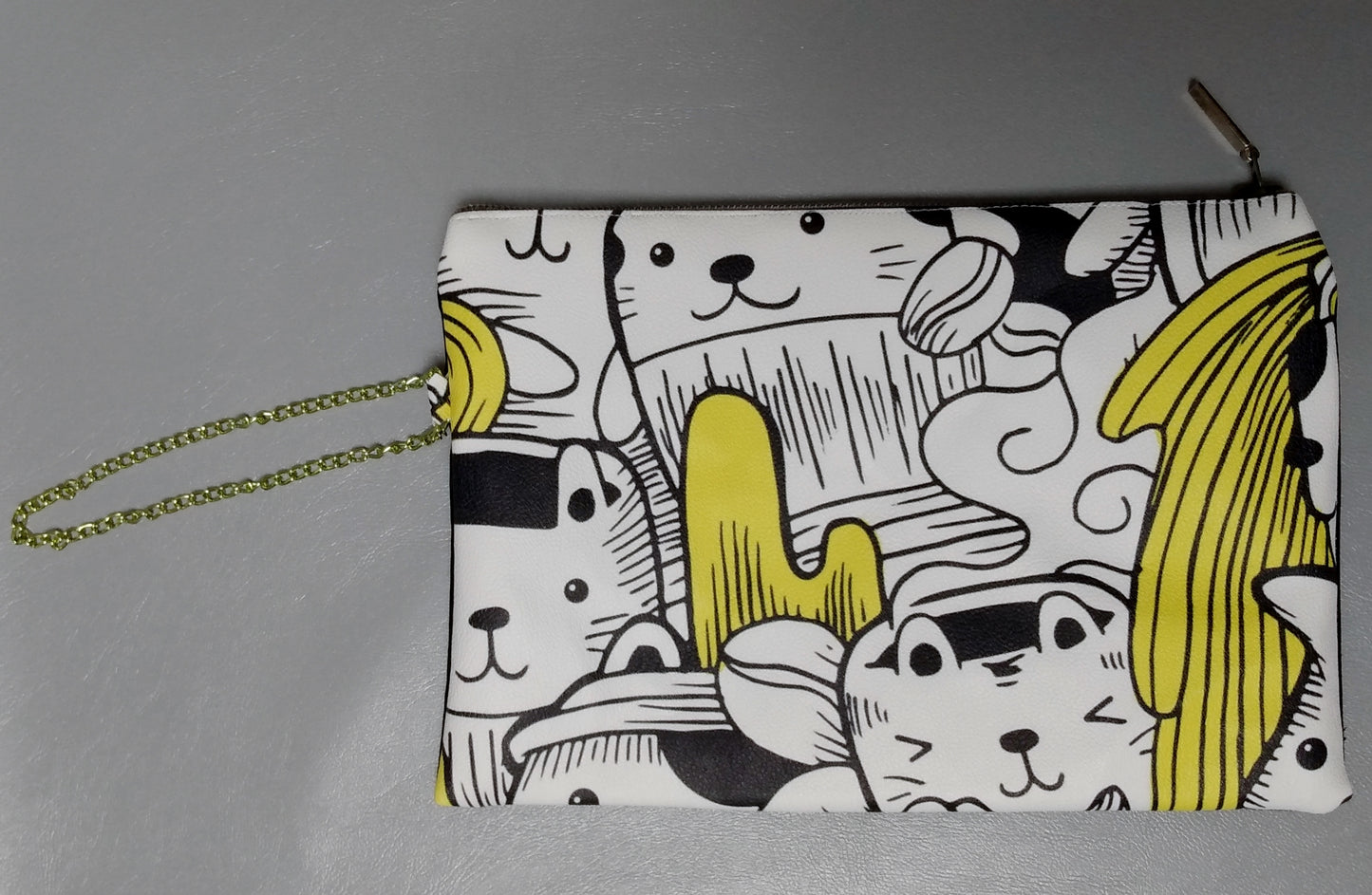 Face avant de la pochette Bonidé sans noeud. Elle est en simili cuir imprimé chat blanc, noir et jaune. On peut voir une dragonne, qui est une chainette couleur or-vert sur le côté. La pochette se ferme avec une fermeture éclair dorée.