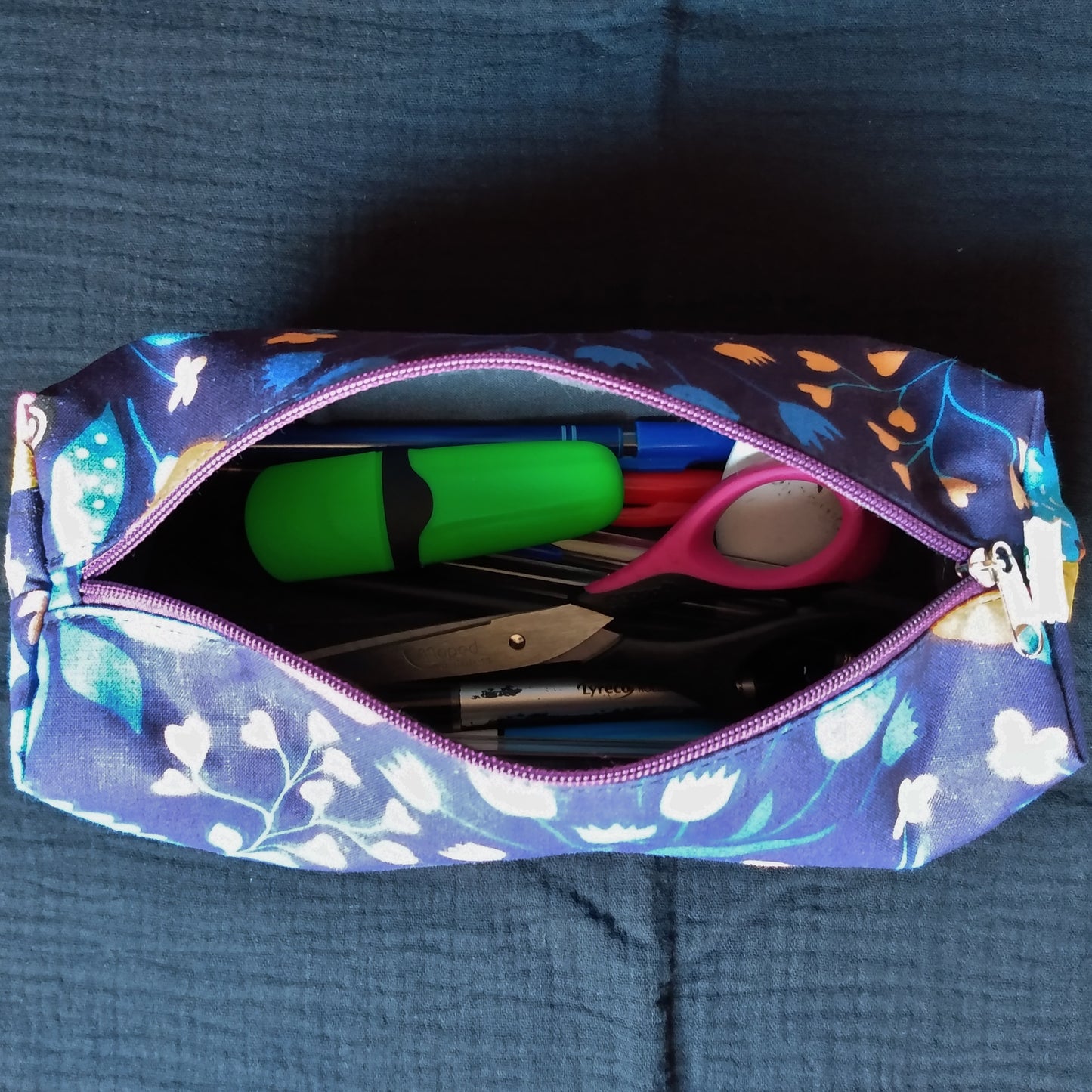 Trousse imprimé fleurs sur fond bleu marine. Elle est ouverte, ce qui laisse entrvoir des fournitures scolaires (ciseaux, stylos...) et la doublure gris anthracite.