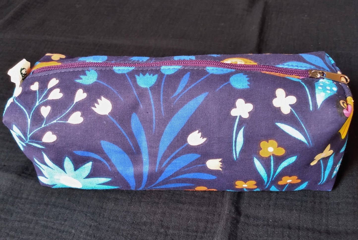 Trousse taille 1 imprimé fleurs sur fond bleu marine. On voit qu'elle se ferme par une fermture éclair violette. L'étiquette "CdN" de la marque est présente sur un côté de la trousse.