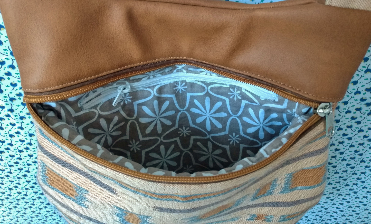 vue avant du sac pivoine marron. Bas du sac en tissu type jacquard beige avec des touches bleutées et marrons. Haut du sac en simili cuir marron. Fermeture via un zip passepoilé, qui est ici ouvert. Il laisse entrevoir l'intérieur qui a une poche zippée. La doublure est en coton couleur taupe avec des motifs fleurs chaya blanches.
