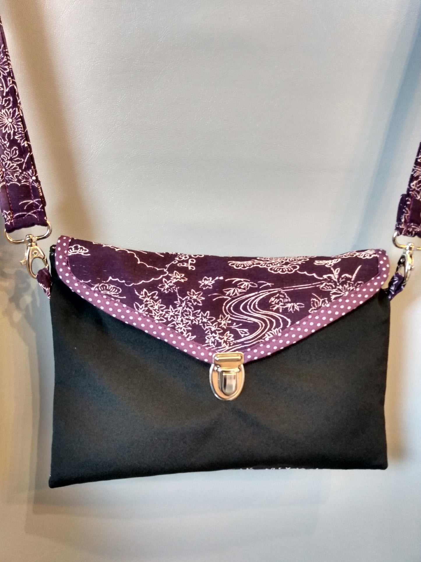 Avant de la pochette Kawa Violet et noir.
Pochette se fermant par un rabat (motifs paysage blanc sur fond violet) et une attache cartable nickelée. Le rabat est entouré d'un biais violet à pois blancs. Le corps du sac est noir. La bandoulière est dans le même tissu que le rabat.