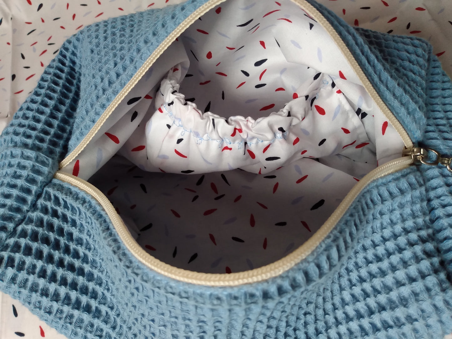 Vue intérieure de la trousse de toilette nid d'abeille bleu clair. La doublure est en coton blanc à motif pépin noir, rouge et bleu. Une poche élastiquée est présente à l'intérieur de la trousse.