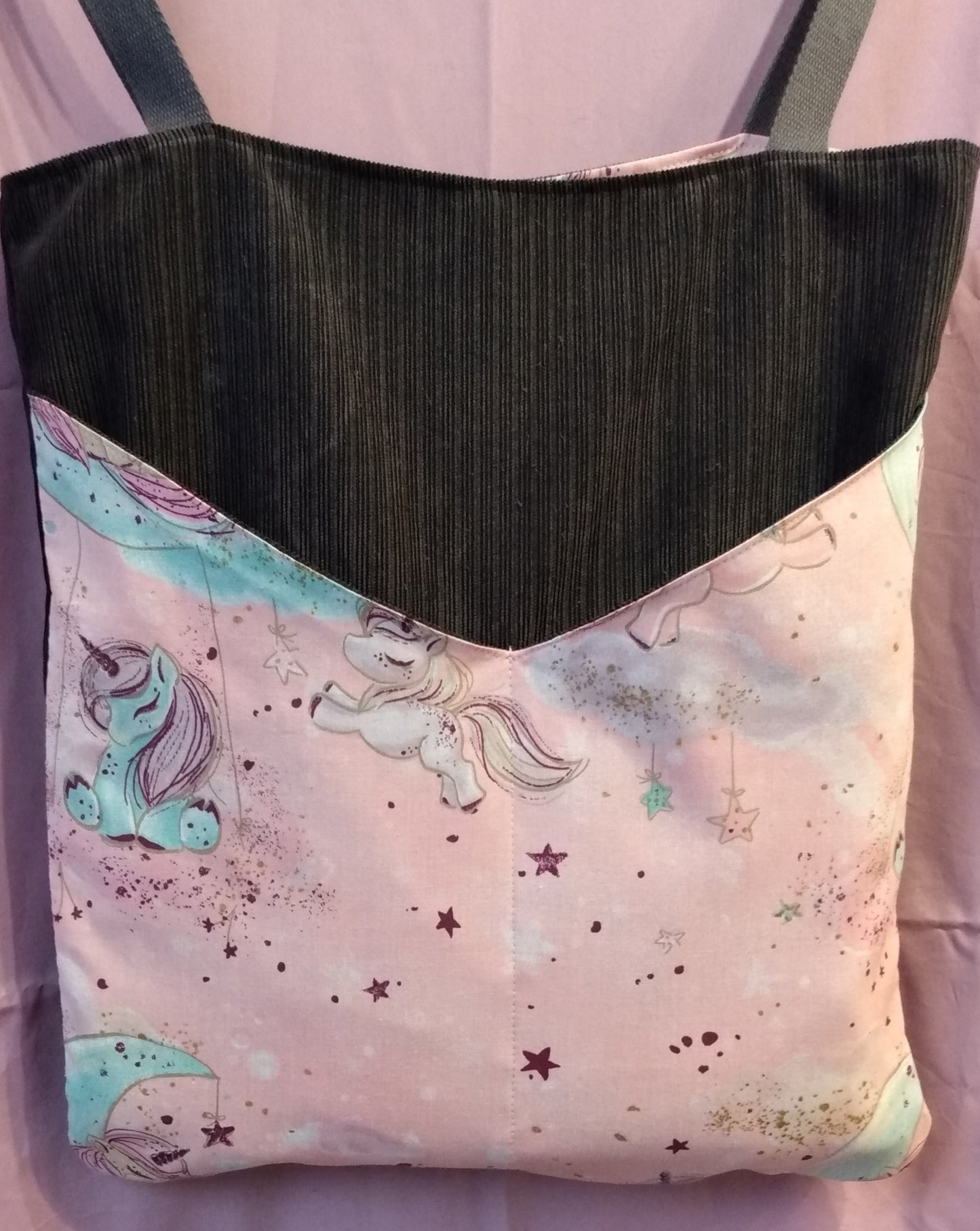 Avant du sac Balad'In rose et gris, tissu principal velours gris anthracite avec deux poches plaquées, qui partent du bas jusqu'aux 3/4 du sac environ. Poches motif licornes et étoiles couleurs pâles. Les anses sont faites pour porter le sac à l'épaule et sont en coton gris.