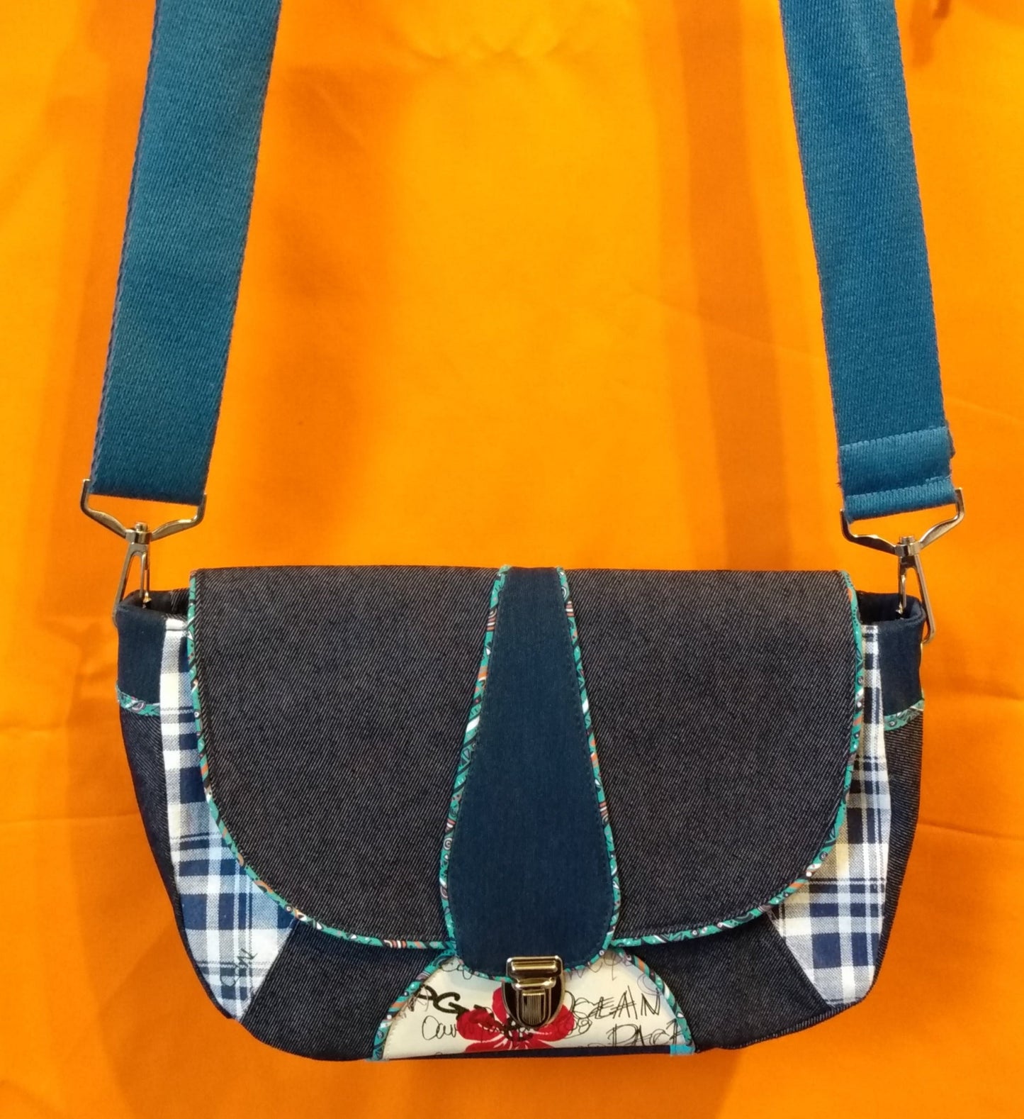 Avant du sac Vegas en jean. Le rabat, en jean bleu foncé et simili cuir bleu foncé au centre avec passepoil turquoise à motifs géométriques, se ferme avec un fermoir cartable. La bandoulière réglable, attachée par deux mousquetons, est en coton bleu. Le corps du sac est en jean à carreaux bleu et blanc avec un triangle jean bleu foncé au centre et un demi cercle en simili cuir beige avec une fleur rouge et des écritures noires où est attachée la base du fermoir.