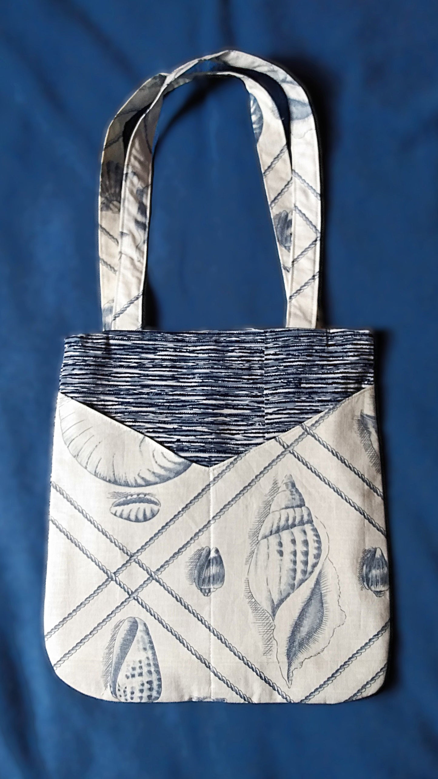 Avant du sac Balad'In coquillages bleus. Le tissu principal est issu de kimono japonais et a des motifs de vaguelettes et lignes horizontales bleu clair, bleu marine et blanches. On voit deux poches plaquées, qui partent du bas jusqu'aux 3/4 du sac environ. Elles sont en coton blanc à motif gros coquillages et cordelettes bleu gris. Les anses sont faites pour porter le sac à l'épaule et sont dans le même tissu que les poches.
