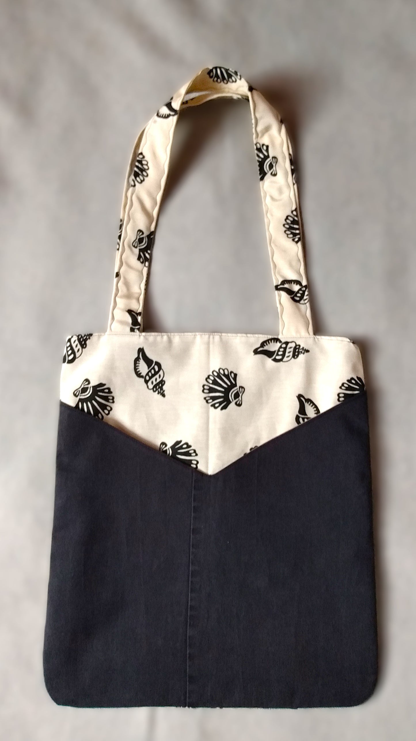Avant du sac Balad'In coquillages noirs et jean noir. Le tissu principal est écru irisé avec motifs gros coquillages noirs. On voit deux poches plaquées, qui partent du bas jusqu'aux 3/4 du sac environ. Poches en jean noir. Les anses sont faites pour porter le sac à l'épaule et sont en tissu principal.
