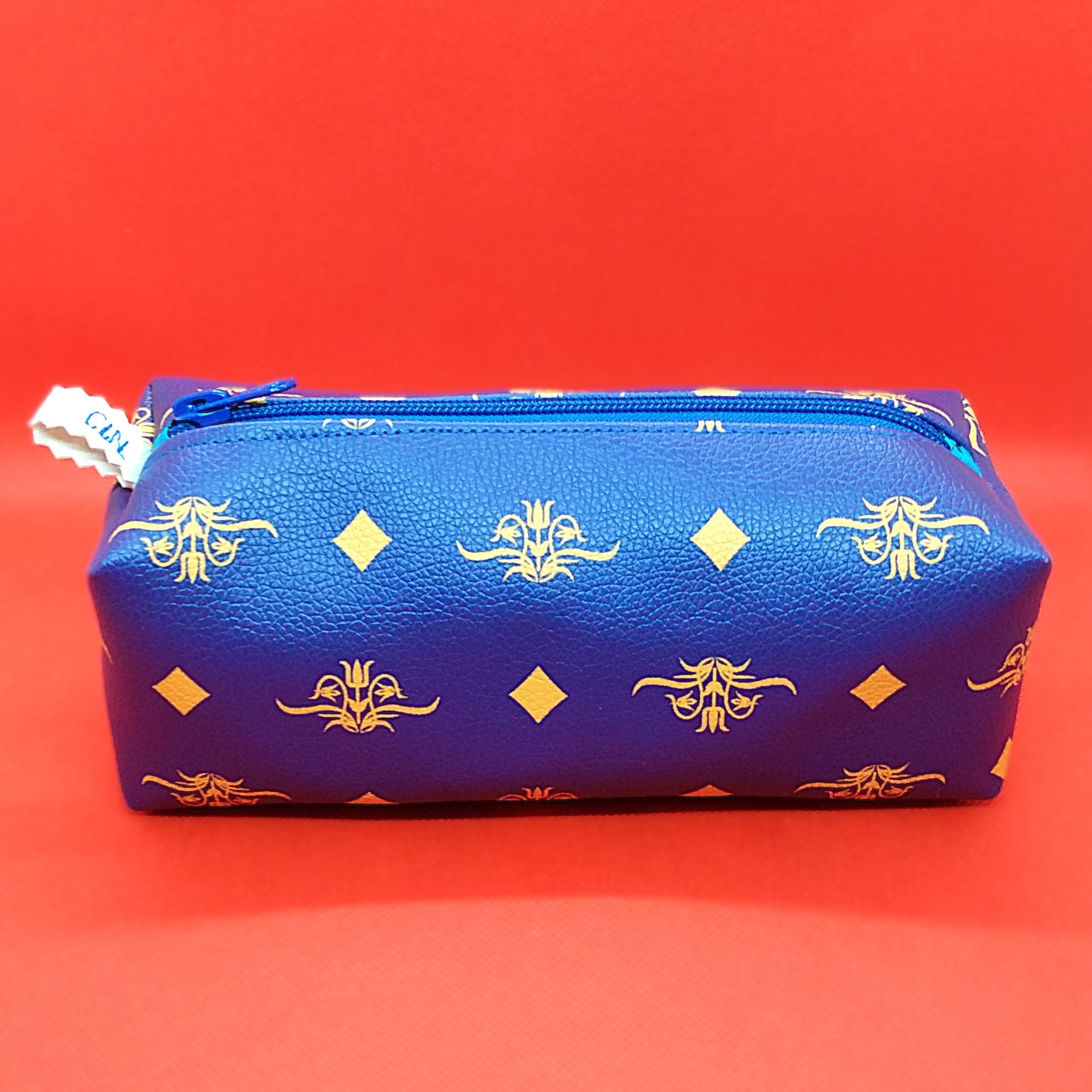 Trousse taille 1 simili cuir bleu roi à fleurs et losanges jaunes. Se ferme par une fermeture éclair bleue. L'étiquette "CdN" de la marque est présente sur un côté de la trousse.