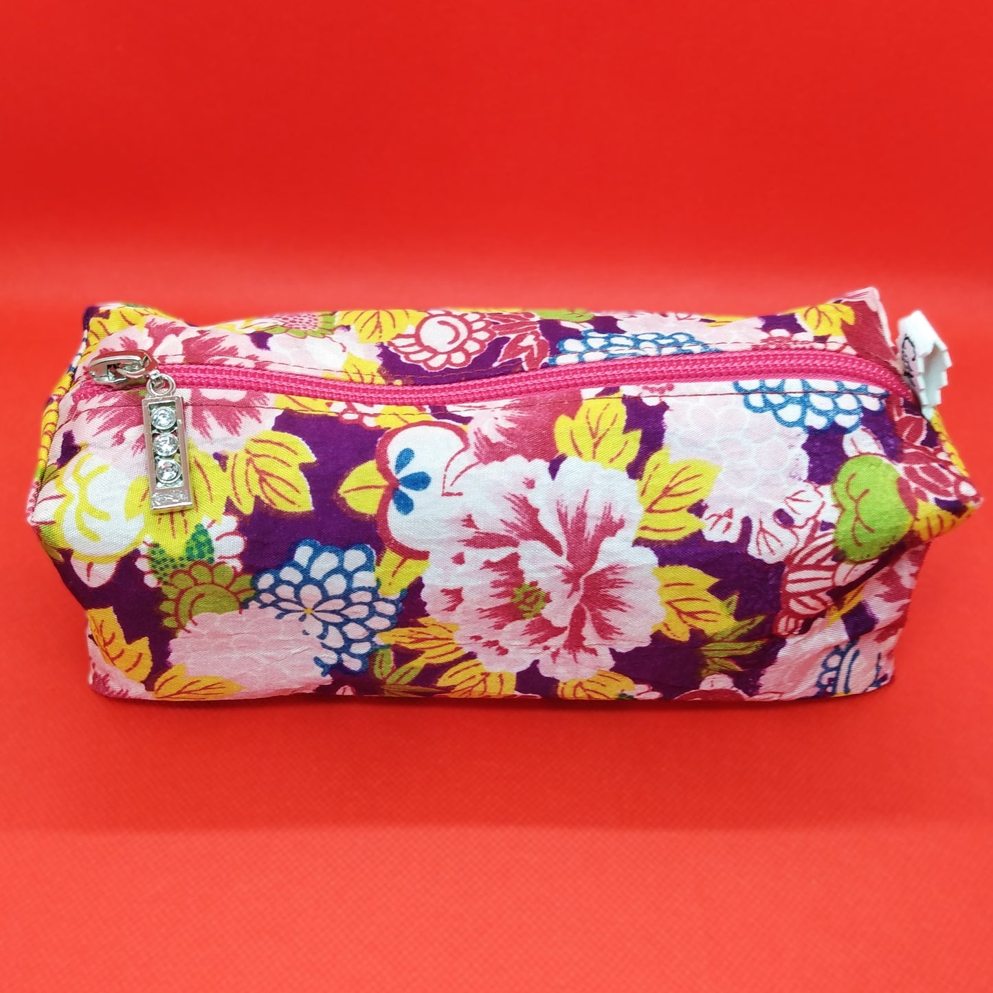 Trousse taille 1 tissu à grosses fleurs roses, jaunes, bleues ou vertes, sur fond violet foncé. Se ferme par une fermeture éclair rose. L'étiquette "CdN" de la marque est présente sur un côté de la trousse.