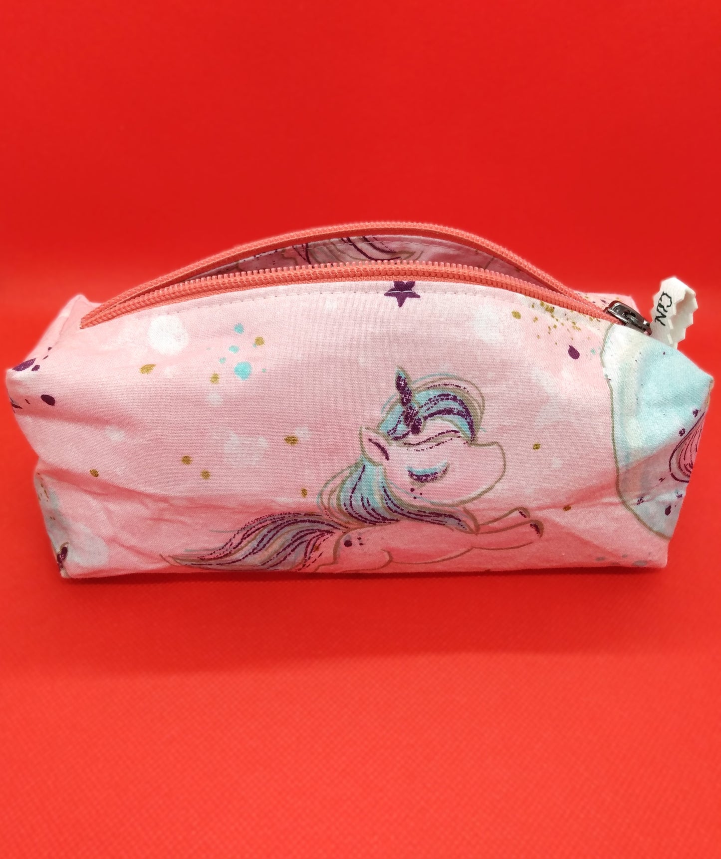 Trousse taille 1 motif grosse licorne et étoiles sur fond rose. Les couleurs sont pastels. Se ferme par une fermeture éclair rose. L'étiquette "CdN" de la marque est présente sur un côté de la trousse.