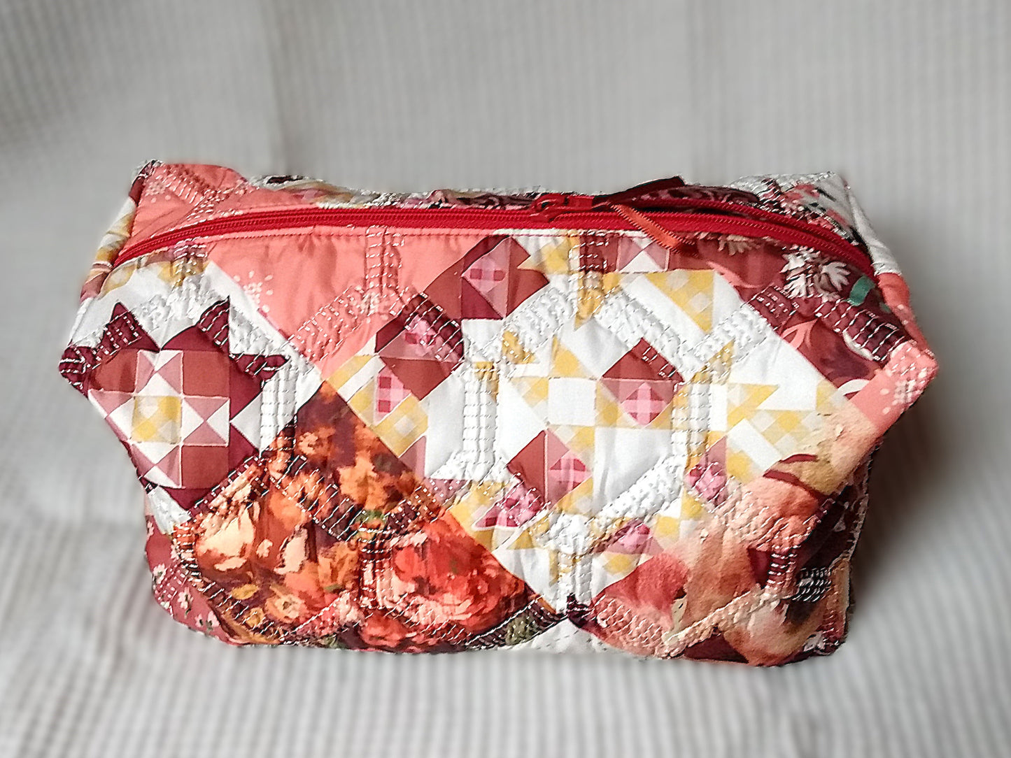 Trousse de toilette Flamingo. Vue de devant. Cette trousse de toilette est rectangulaire, elle se ferme avec une fermeture éclair rouge. L'extérieur est en tissu matelassé imprimé en patchwork dans les tons roses, bordeaux, blanc, fleuri...