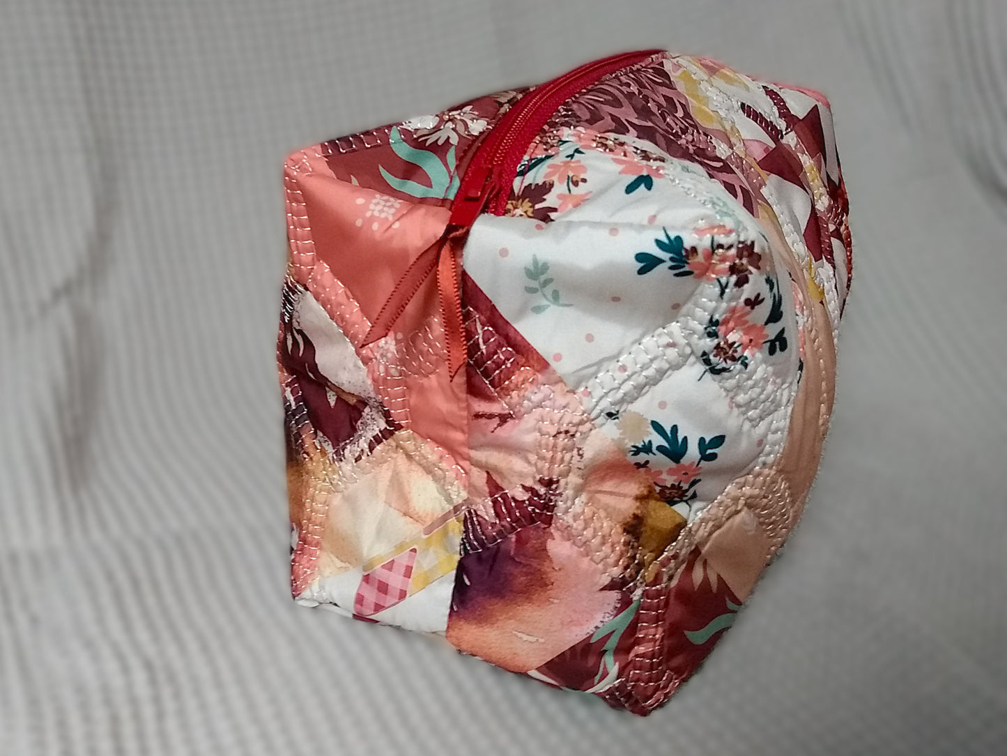 Trousse de toilette Flamingo. Vue de côté. Cette trousse de toilette est rectangulaire, elle se ferme avec une fermeture éclair rouge. L'extérieur est en tissu matelassé imprimé en patchwork dans les tons roses, bordeaux, blanc, fleuri...