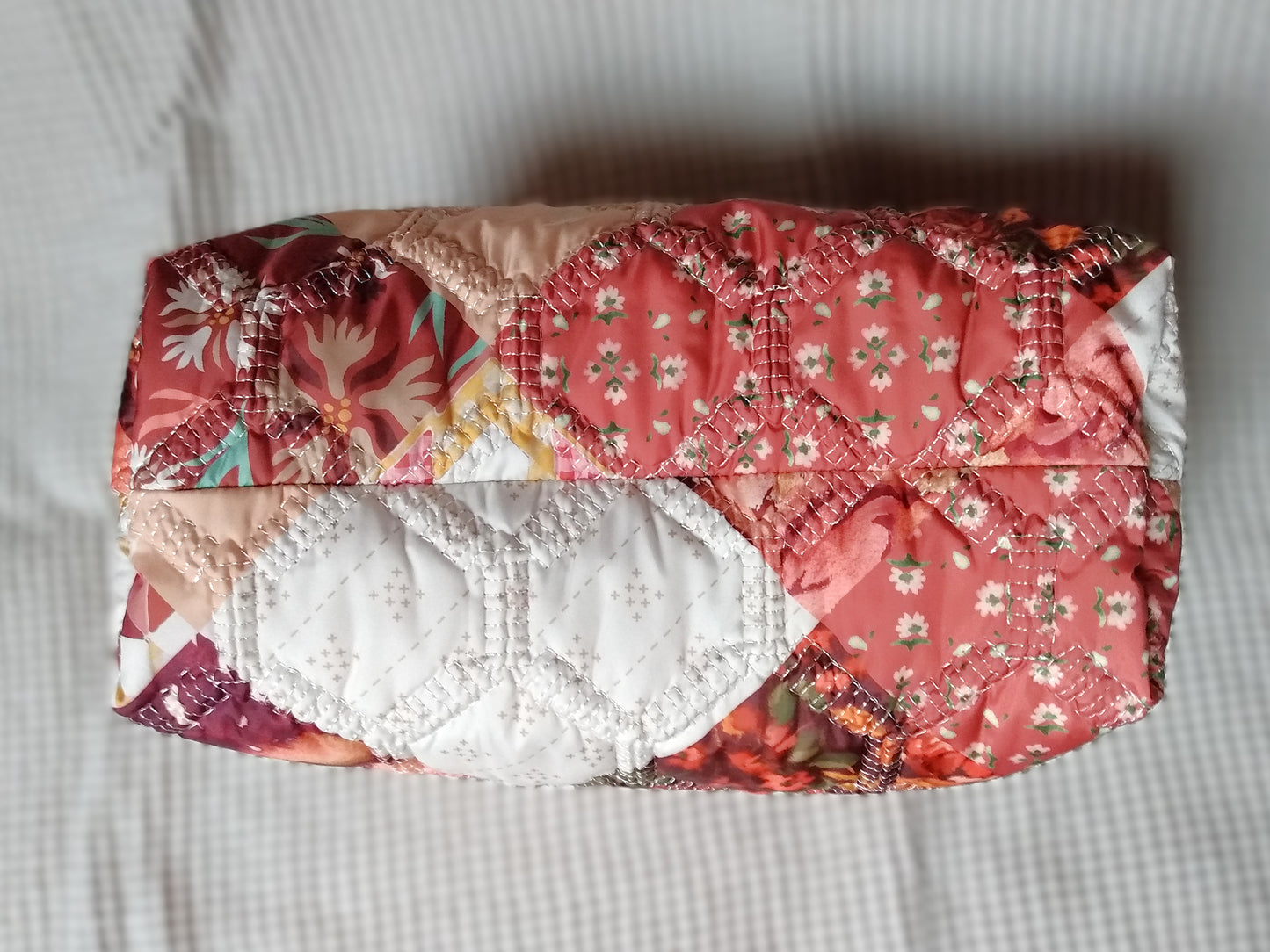 Trousse de toilette Flamingo. Vue du dessous. Cette trousse de toilette est rectangulaire, elle se ferme avec une fermeture éclair rouge. L'extérieur est en tissu matelassé imprimé en patchwork dans les tons roses, bordeaux, blanc, fleuri...