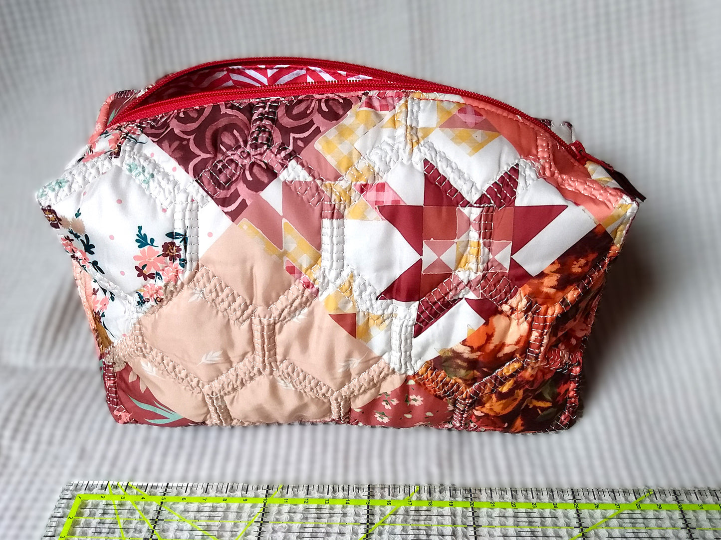 Trousse de toilette Flamingo. Vue de devant. Cette trousse de toilette est rectangulaire, elle se ferme avec une fermeture éclair rouge. L'extérieur est en tissu matelassé imprimé en patchwork dans les tons roses, bordeaux, blanc, fleuri...La trousse ouverte laisse apparaitre la doublure intérieure en tissu à rayures rouges et blanches en chevron.