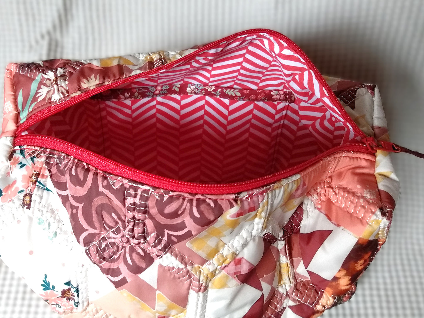 Trousse de toilette Flamingo. Vue intérieure. La trousse de toilette ouverte laisse apparaitre la doublure intérieure en tissu à rayures rouges et blanches en chevron. Une poche plaquée est visible sur un côté de la doublure. Cette poche est surmontée d'un biais bordeaux à fleurs.
