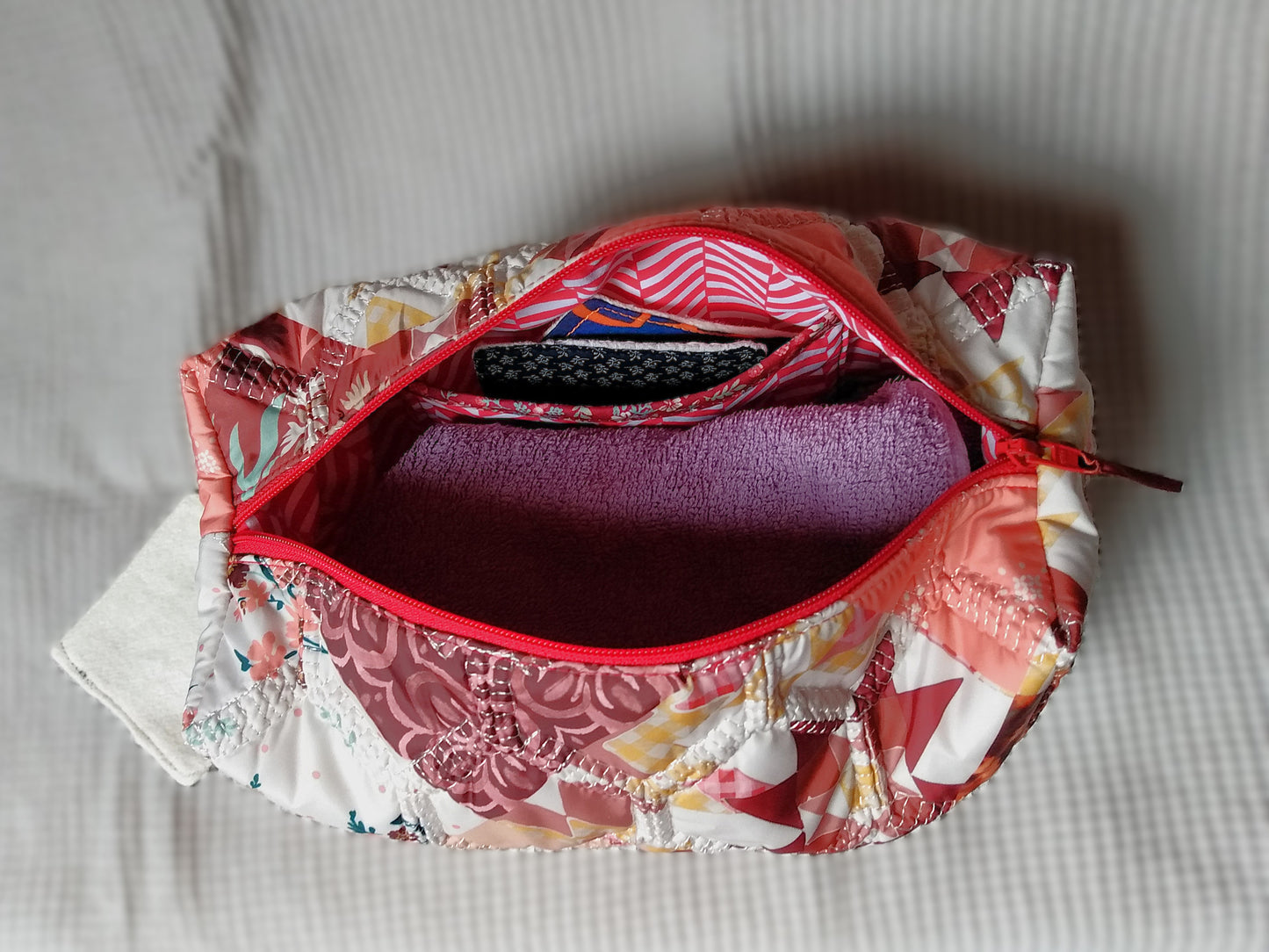 Trousse de toilette Flamingo. Vue intérieure. Cette trousse de toilette est rectangulaire, elle se ferme avec une fermeture éclair rouge. L'extérieur est en tissu matelassé imprimé en patchwork dans les tons roses, bordeaux, blanc, fleuri...La trousse ouverte laisse apparaitre la doublure intérieure en tissu à rayures rouges et blanches en chevron. On peut voir à l'intérieur, une petite serviette de toilette ainsi que des lingettes démaquillantes.