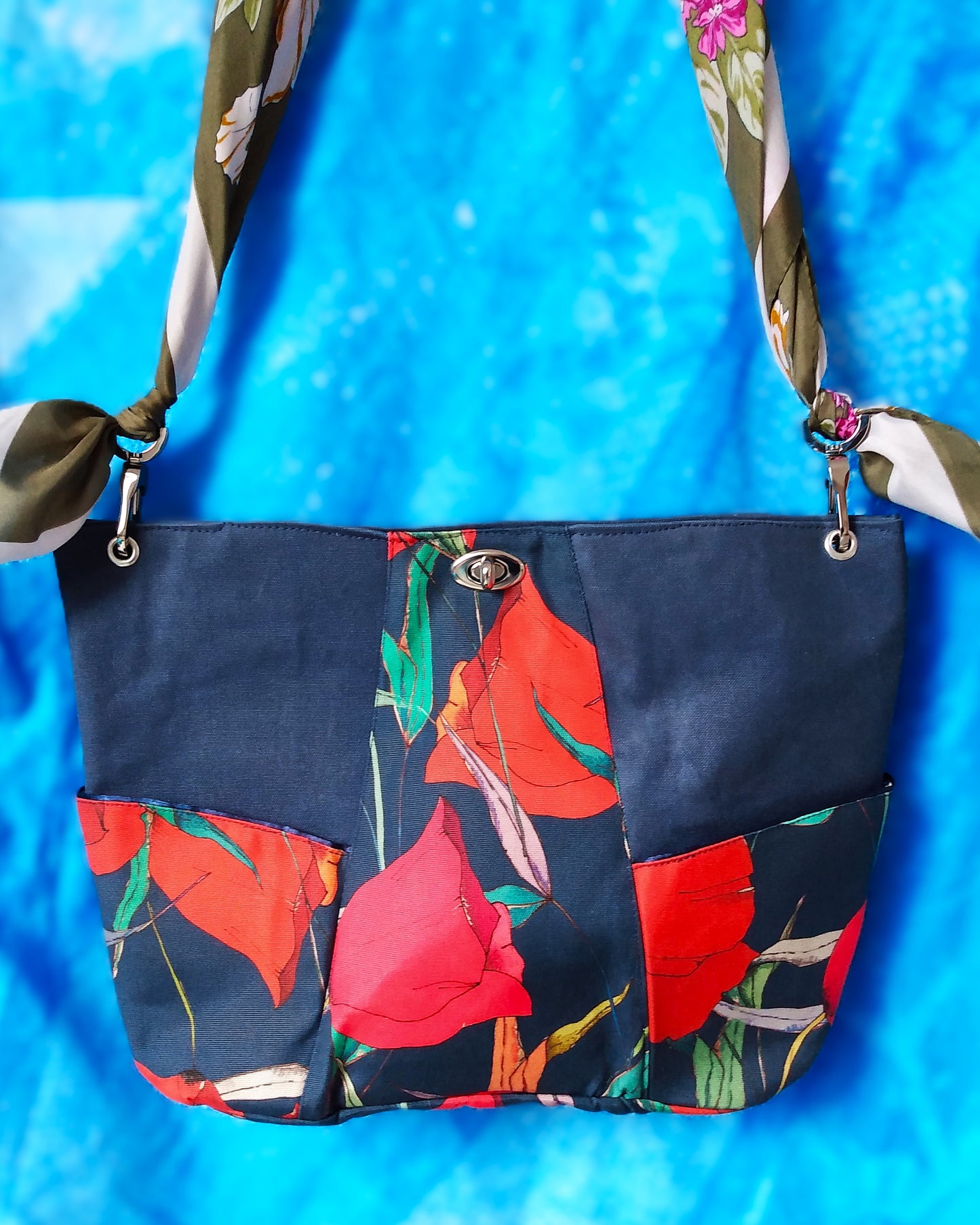 Vue avant Sac capri fleuri. Le corps du sac est composé de 3 bandes verticales. Au centre une bande en tissu déperlant avec imprimé gros coquelicots rouges et feuilles vertes sur fond bleu foncé. Les 2 bandes latérales sont en tissu épais imperméabilisé bleu foncé. 2 poches latérales sont dans le même tissu qu'au centre. Un fermoir tourniquet nickelé ferme le sac. L'anse est un foulard vert kaki et blanc à fleurs roses et blanches. Elle est liée au sac via des mousquetons et œillets nickelés.