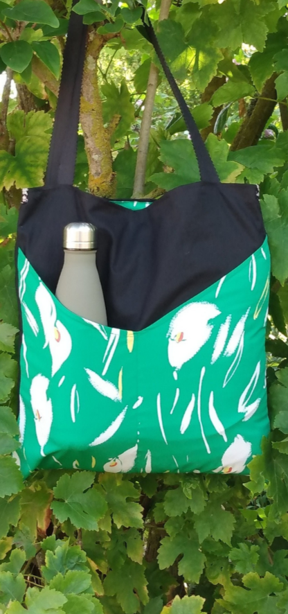 Avant du sac balad'In noir et vert. tissu principal noir et deux poches plaquées vertes avec des fleurs blanches. Anses pour porter le sac à l'épaule en toile à sac noire.