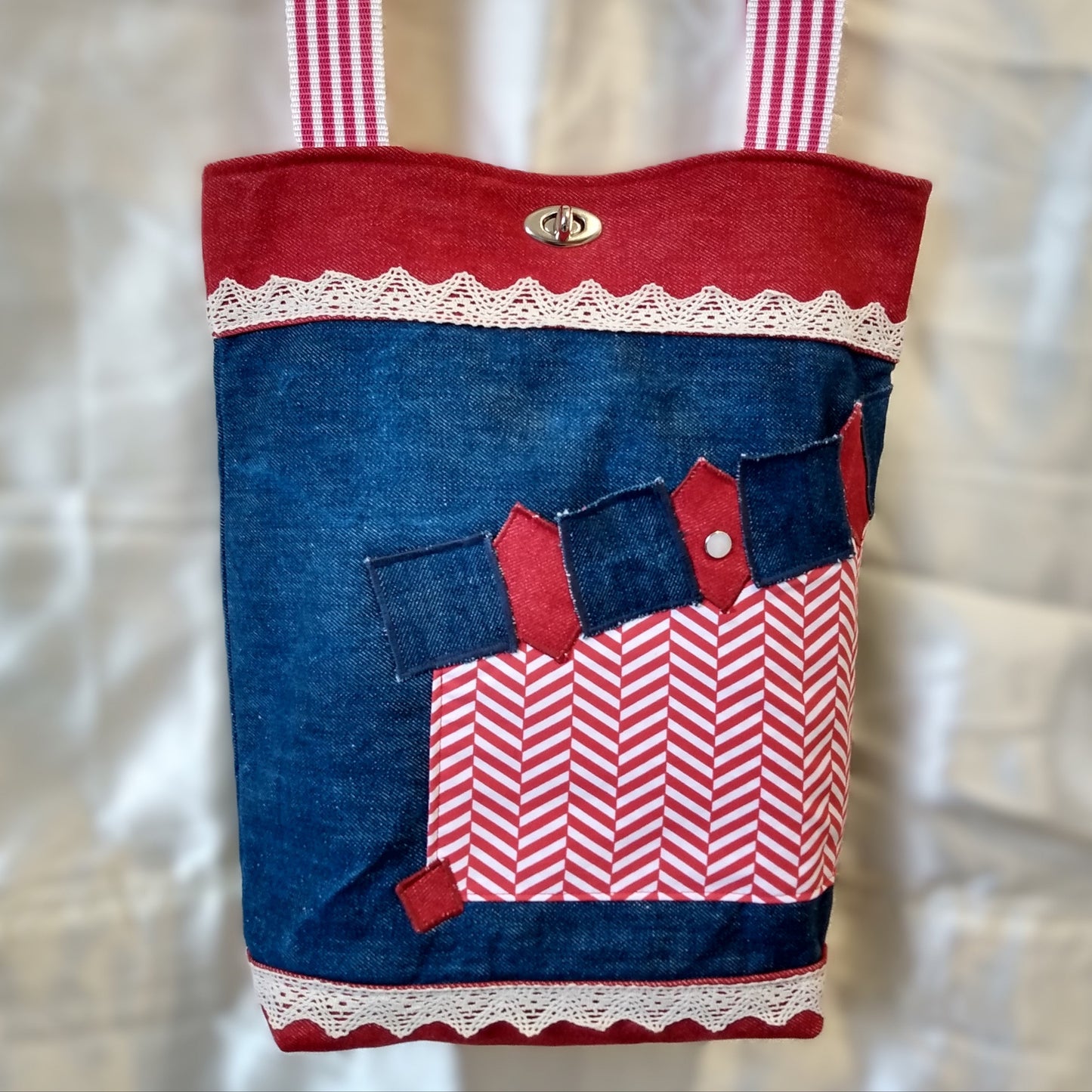 Avant du sac Boujou. Extérieur en denim avec une poche se fermant par un bouton. Fermeture du sac avec un fermoir tourniquet en haut au centre. Anses à rayures rouge et blanche. Bande horizontales en jean rouge en haut (8 cm) et en bas du sac (2 cm). Centre du sac en jean bleu avec une ligne horizontale de dentelle (2cm) en haut et une en bas.
Poche en tissu déperlant à chevrons rouge et blanc, côté gauche du sac. Haut de la poche, alternance de carrés bleus et losanges rouges en jean.