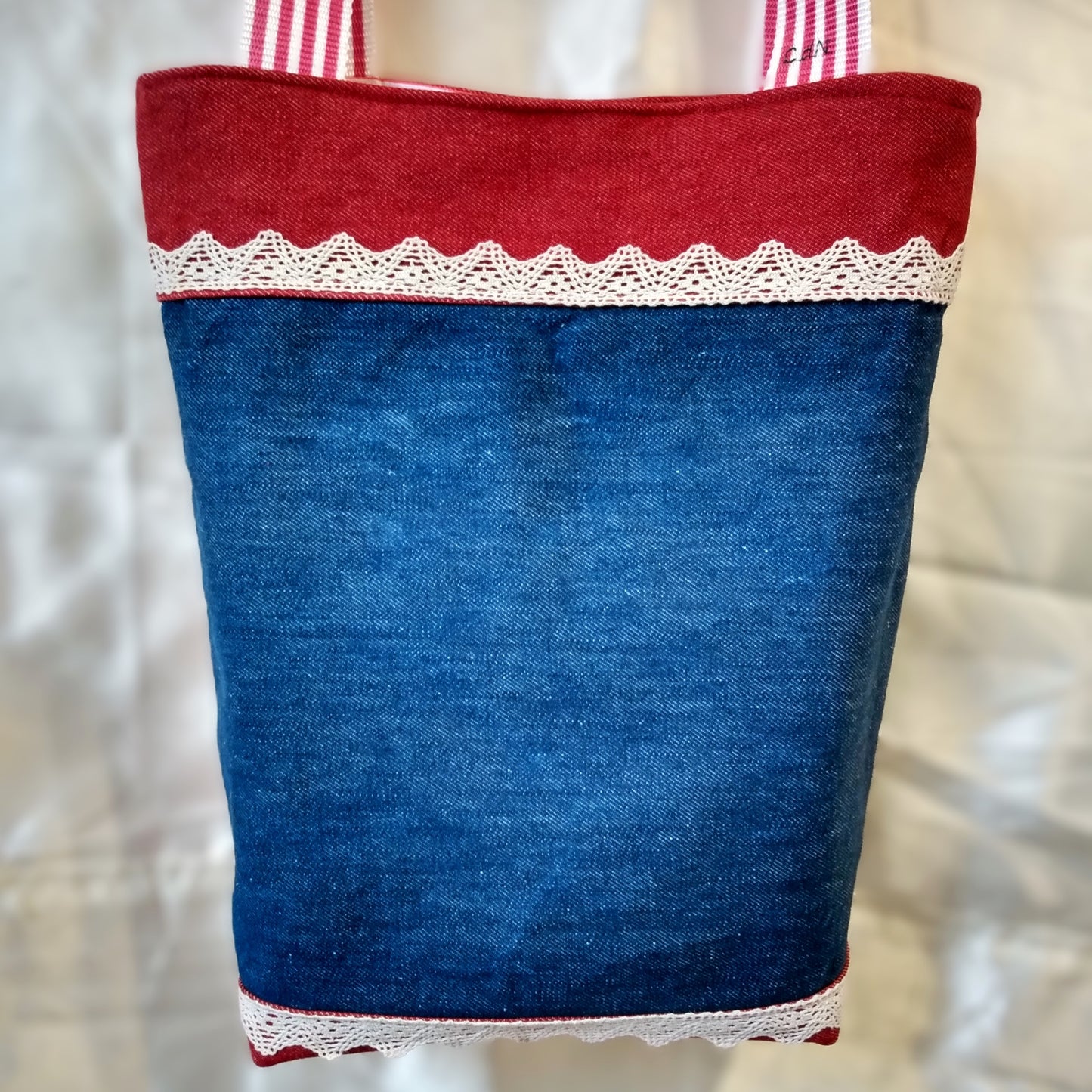 Dos du sac Boujou. Extérieur en denim. Anses à rayures rouge et blanche. Bande horizontales en jean rouge en haut (8 cm) et en bas du sac (2 cm). Centre du sac en jean bleu avec une ligne horizontale de dentelle (2cm) en haut et une en bas.