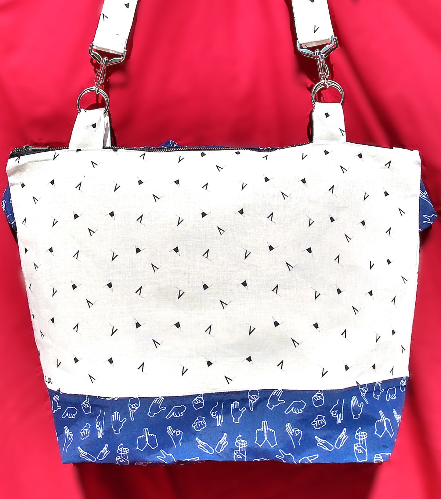 arrière du sac signora. Le corps du sac est en lin blanc à motifs fanions triangulaires bleu marine. Le bas du sac est en coton bleu foncé à motifs mains blanches parlant en langue des signes. Les anses, même tissu que le corps du sac, sont accrochées via des anneaux et des mousquetons. Le sac se ferme avec une fermeture éclair.
