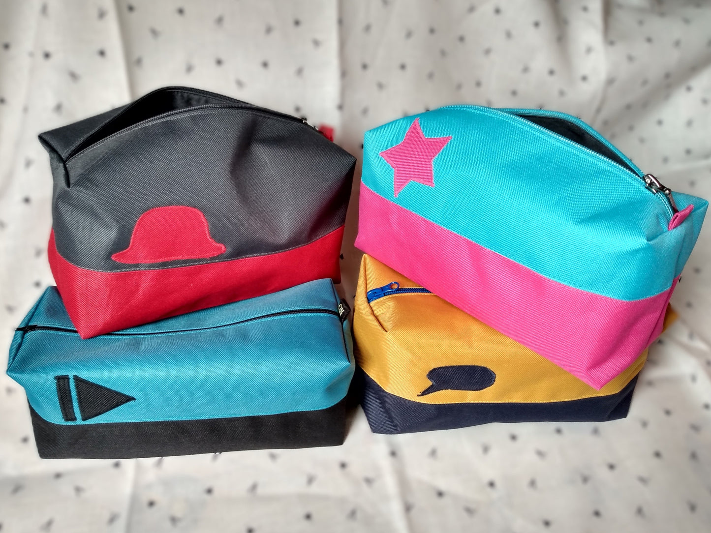 Quatre trousses de toilette Corail. Elles sont rectangulaires et bicolores, en horizontal. 1 noir en bas et bleu paon en haut, avec un appliqué "suivant" noir sur le bleu. 2 bleu foncé en bas et jaune moutarde en haut avec un appliqué bulle de bande dessinée bleu sur le jaune. 3 rouge en bas et gris anthracite en haut avec un chapeau melon rouge sur le gris. 4 rose en bas et bleu ciel en haut avec une étoile rose sur le bleu. Elles se ferment avec une fermeture éclair.