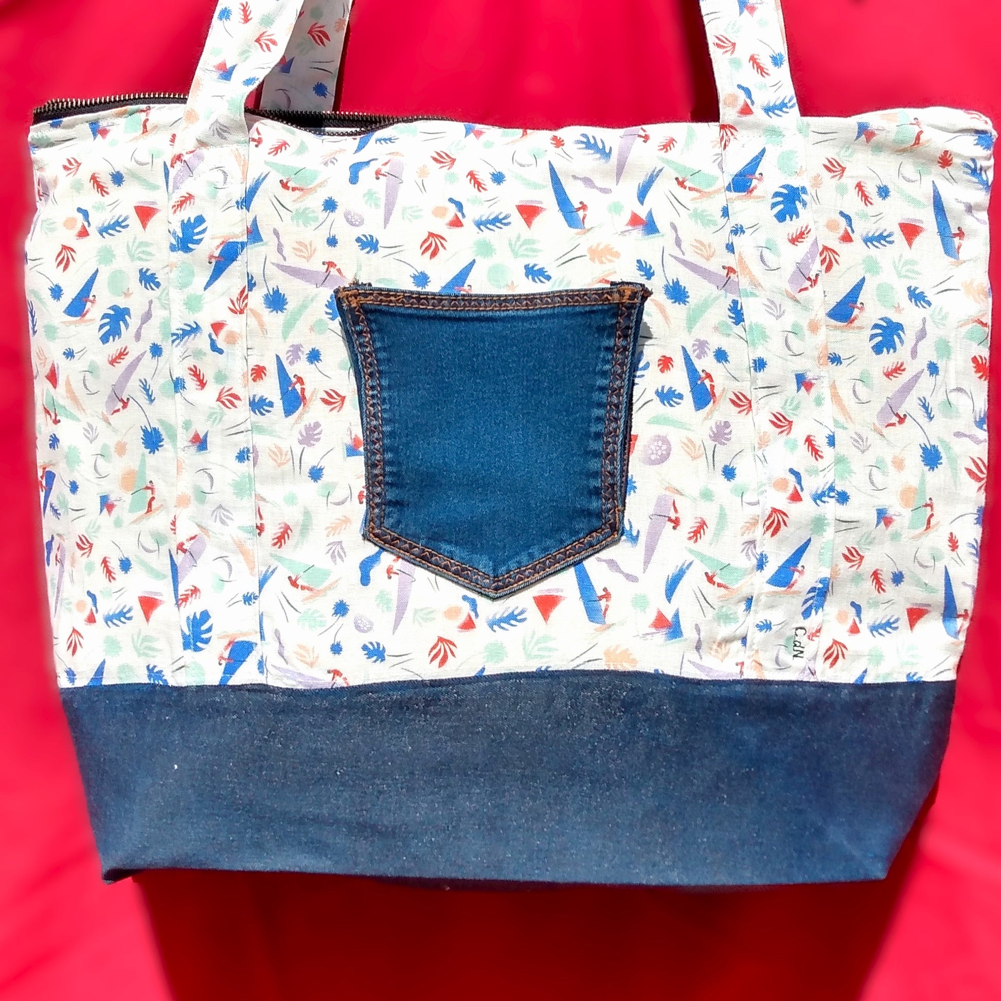Arrière du sac Bordemer. Le corps du sac est en lin beige à petits motifs feuilles et planches à voile de plusieurs couleurs plutôt pastel. Au centre, une poche arrière de jean sert de poche plaquée. Le bas du sac est en jean. Les anses sont dans le même tissu que le corps du sac. Le sac se ferme avec une fermeture éclair métal.