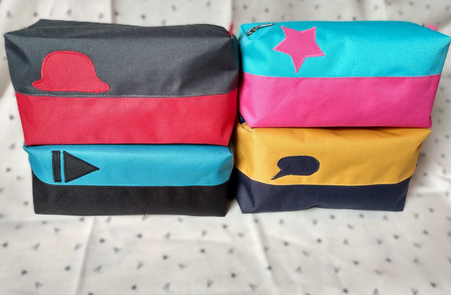 Quatre trousses de toilette Corail. Elles sont rectangulaires et bicolores, en horizontal. 1 noir en bas et bleu paon en haut, avec un appliqué "suivant" noir sur le bleu. 2 bleu foncé en bas et jaune moutarde en haut avec un appliqué bulle de bande dessinée bleu sur le jaune. 3 rouge en bas et gris anthracite en haut avec un chapeau melon rouge sur le gris. 4 rose en bas et bleu ciel en haut avec une étoile rose sur le bleu. Elles se ferment avec une fermeture éclair.