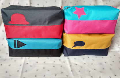 Quatre trousses de toilette Corail. Elles sont rectangulaires et bicolores, en horizontal. 1 noir en bas et bleu paon en haut, avec un appliqué "suivant" noir sur le bleu. 2 bleu foncé en bas et jaune moutarde en haut avec un appliqué bulle de bande dessinée bleu sur le jaune. 3 rouge en bas et gris anthracite en haut avec un chapeau melon rouge sur le gris. 4 rose en bas et bleu ciel en haut avec une étoile rose sur le bleu. Elles se ferment avec une fermeture éclair.