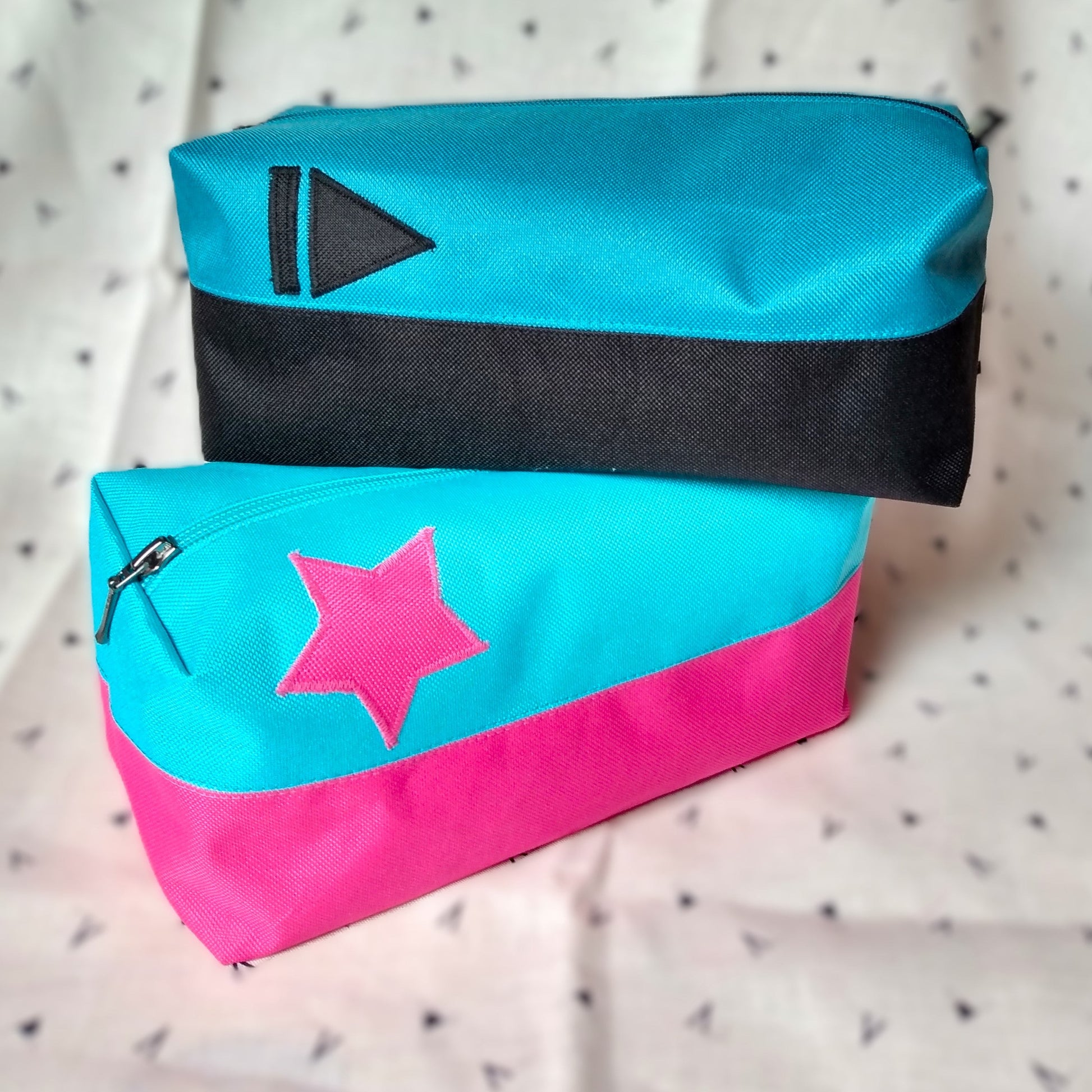 Deux trousses de toilette Corail qui se chevauchent. Elles sont rectangulaires et bicolores, en horizontal. L'une est noire en bas et bleu paon en haut, avec un appliqué "suivant" (trait vertical et triangle) noir sur le bleu. La seconde est rose en bas et bleu ciel en haut avec un appliqué en forme d'étoile rose sur le bleu. Elles se ferment avec une fermeture éclair.
