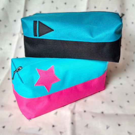 Deux trousses de toilette Corail qui se chevauchent. Elles sont rectangulaires et bicolores, en horizontal. L'une est noire en bas et bleu paon en haut, avec un appliqué "suivant" (trait vertical et triangle) noir sur le bleu. La seconde est rose en bas et bleu ciel en haut avec un appliqué en forme d'étoile rose sur le bleu. Elles se ferment avec une fermeture éclair.