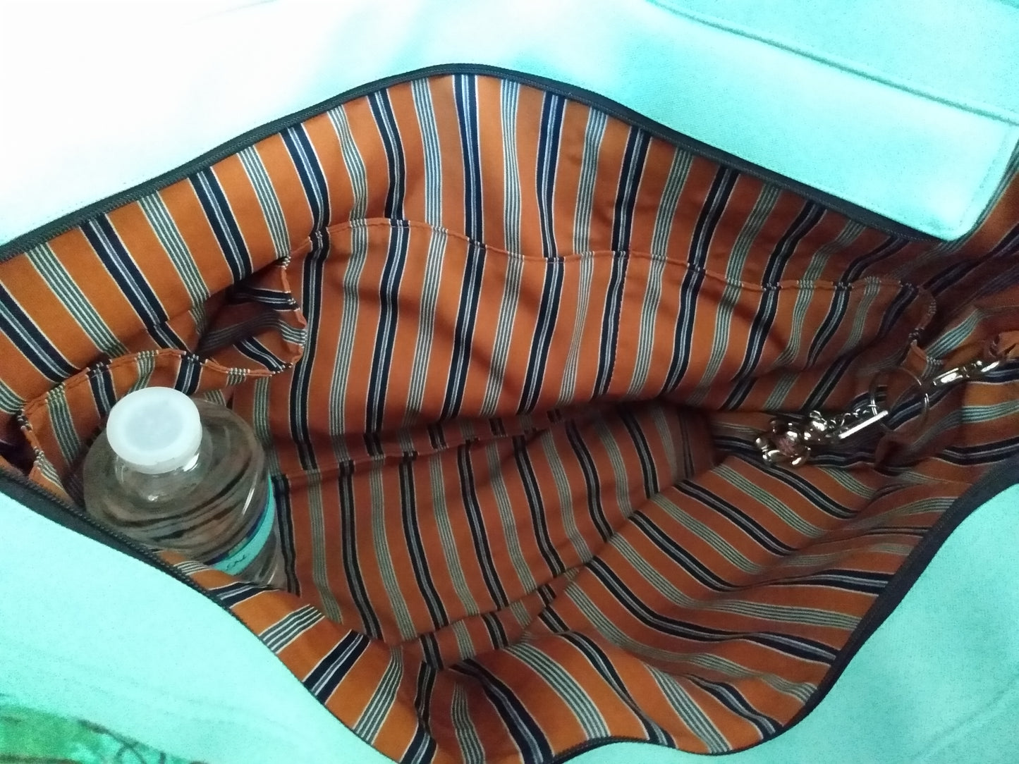 Vue de l'intérieur du sac cabas Amarante. La doublure est de couleur orangée rayée marine. L'intérieur possède une grande poche zippée, 3 poches  ouvertes, un élastique pour tenir une gourde et un mousqueton où sont accrochées des clés.