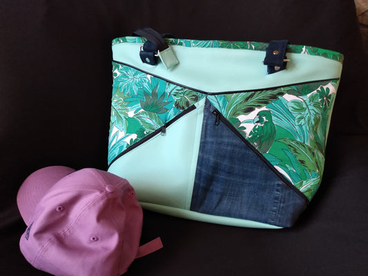 Vue de l'avant du sac cabas Amarante. Sac dans les coloris bleu et vert. Il est découpé en plusieurs parties triangulaires cousues dans différents tissus : simili cuir vert d'eau, coton imprimé feuilles vertes et oiseau, jean.  Il possède deux poches zippées à l'avant. et deux anses accrochées par une boucle rectangulaire et des rivets.