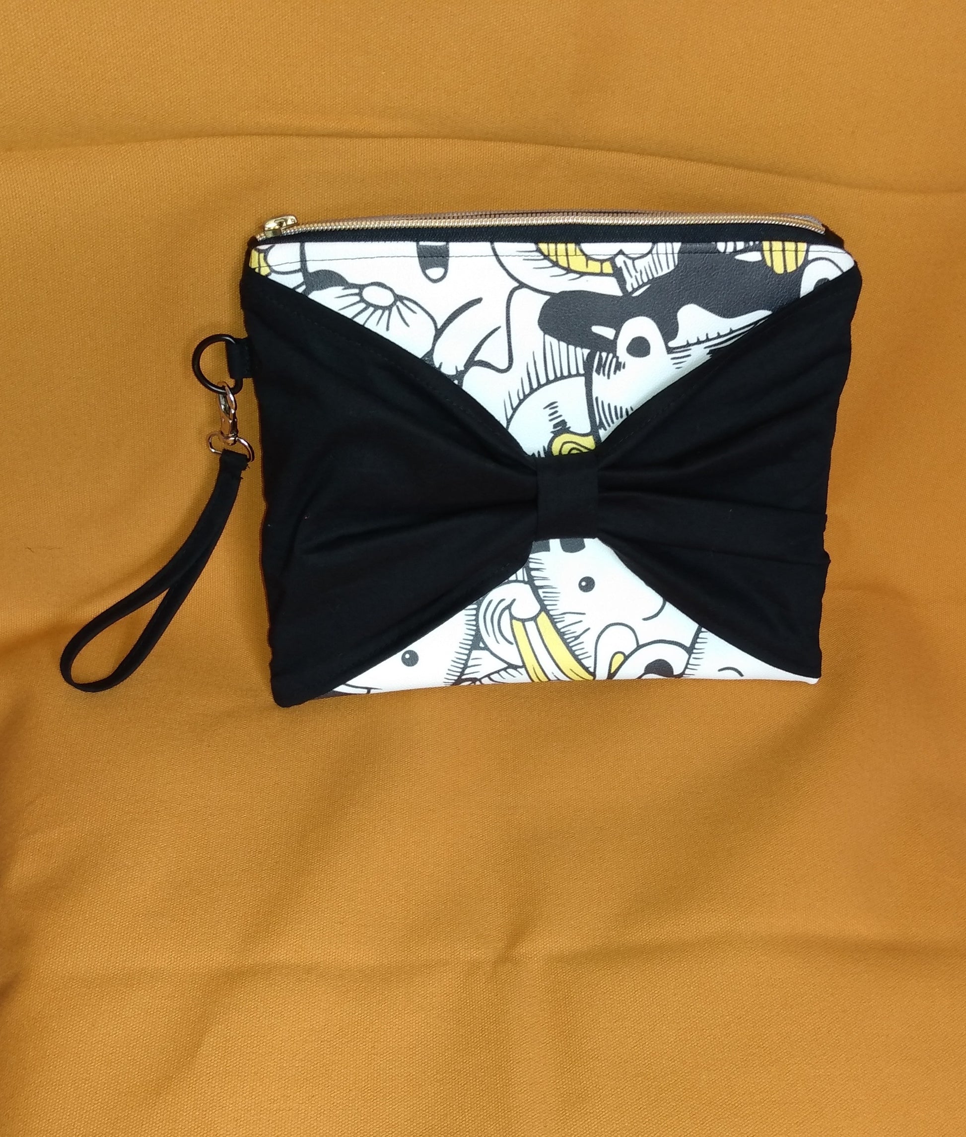 Face avant de la pochette Bonidé. Elle est en simili cuir imprimé chat blanc, noir et jaune. Un noeud est cousu sur l'avant et cache une partie de la face. Une dragonne est visible sur le côté, accrochée avec un mousqueton. La pochette se ferme en haut avec une fermeture éclair dorée.