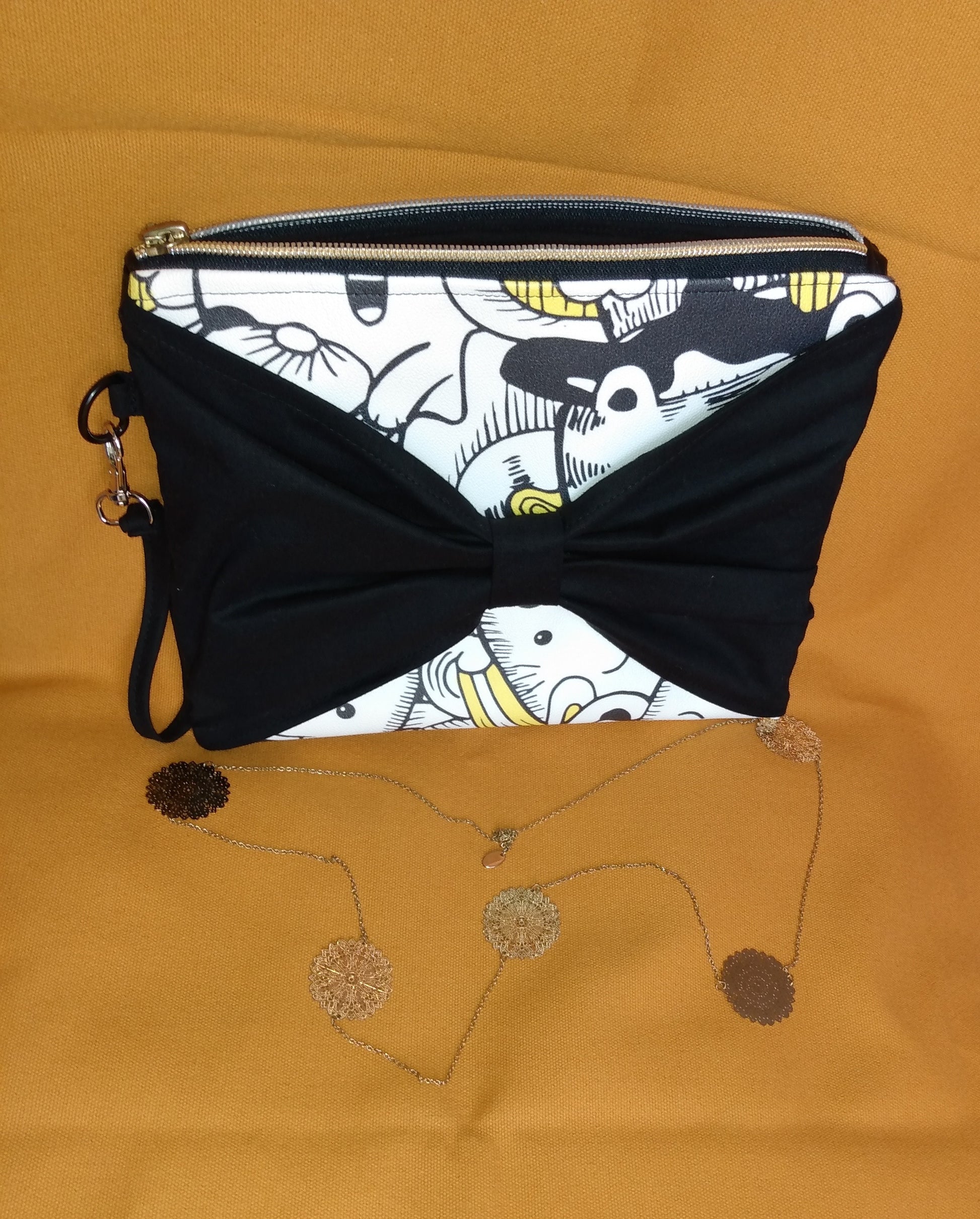 Face avant de la pochette Bonidé. Elle est en simili cuir imprimé chat blanc noir et jaune. Un noeud papillon est en avant et cache donc une partie de la face. On peut voir une dragonne noire sur le côté, accrochée par un mousqueton. La pochette se ferme avec une fermeture éclair dorée.