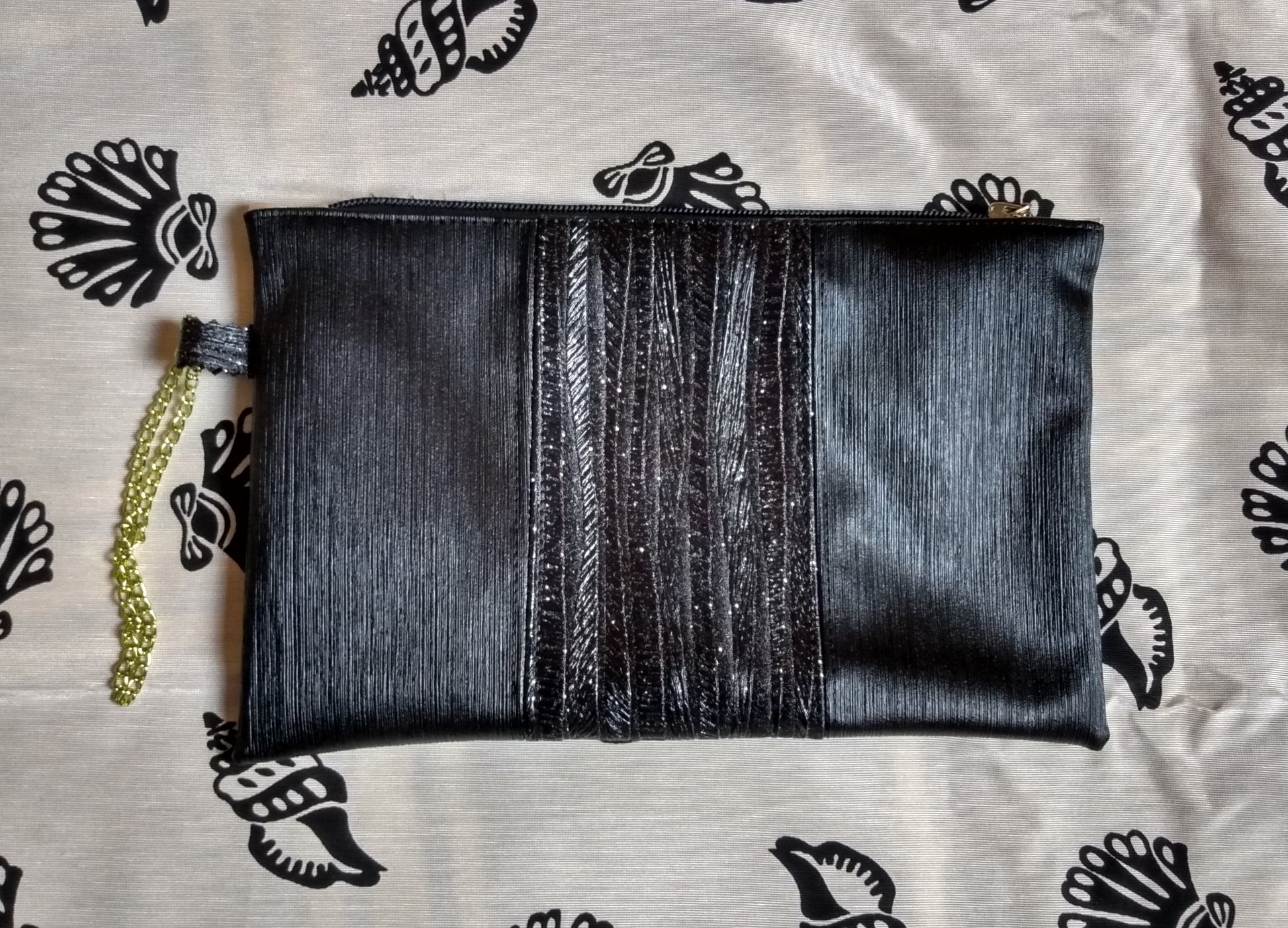 Vue avant pochette Soglad. 3 bandes verticales noires. La bande du milieu contient des paillettes. Une dragonne en chainette permet le port de la pochette au poignet. Une fermeture éclair ferme la pochette.