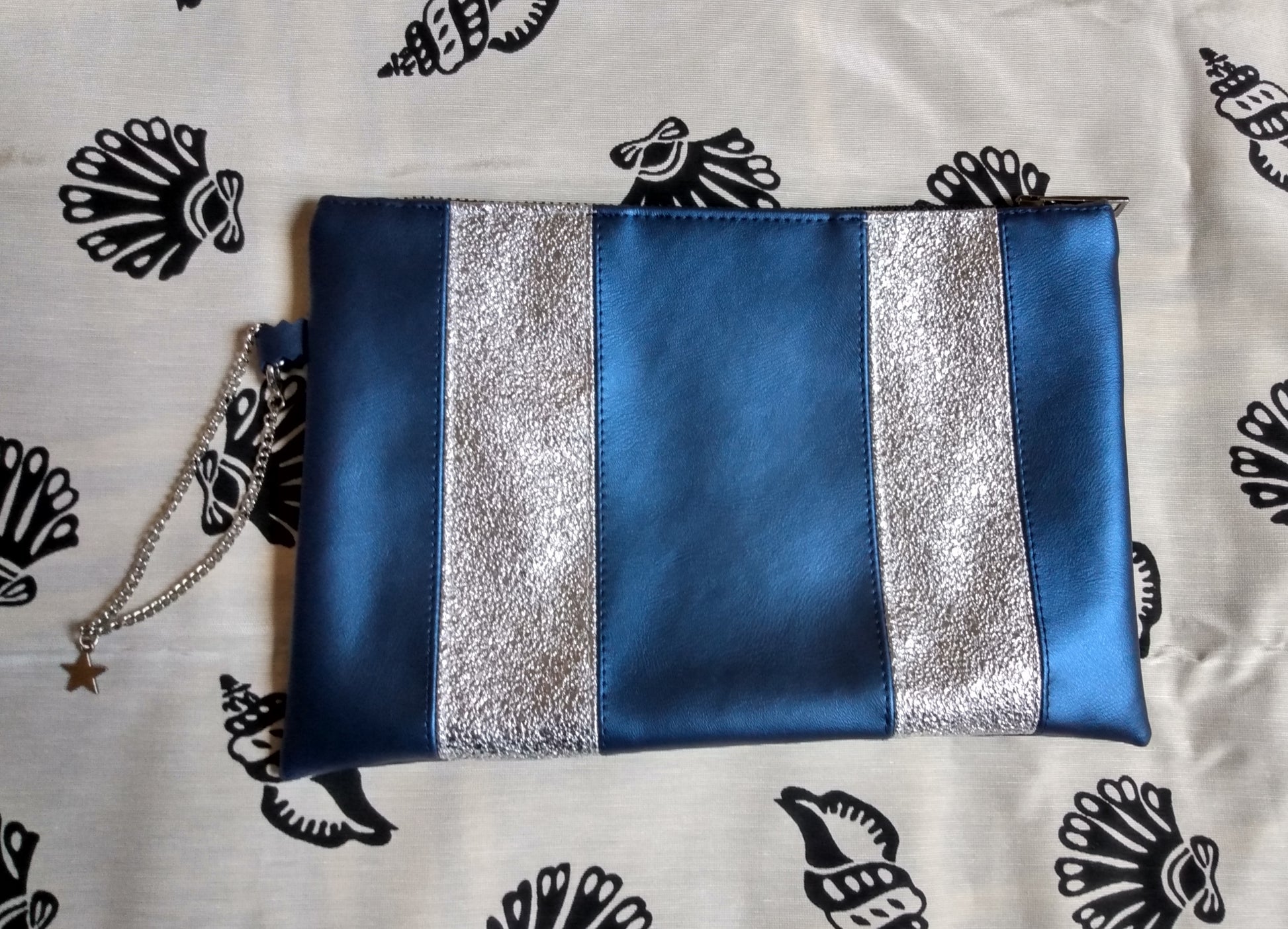 Vue avant pochette Soglad bleu et argent. Des bandes verticales (5) alternes entre bleu métallisé et argent. une chainette permet le port au poignet. Une fermeture éclair ferme la pochette.