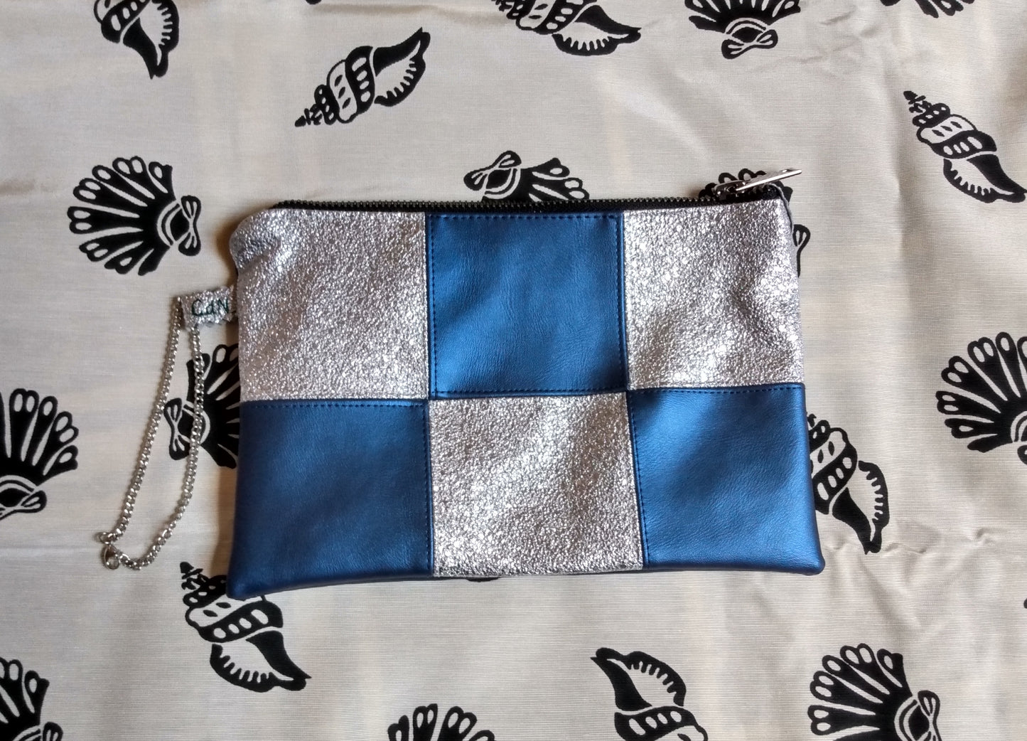 vue avant pochette formidam. Damier bleu et argent. Dragonne sur le côté. Fermeture éclair pour fermer la pochette.