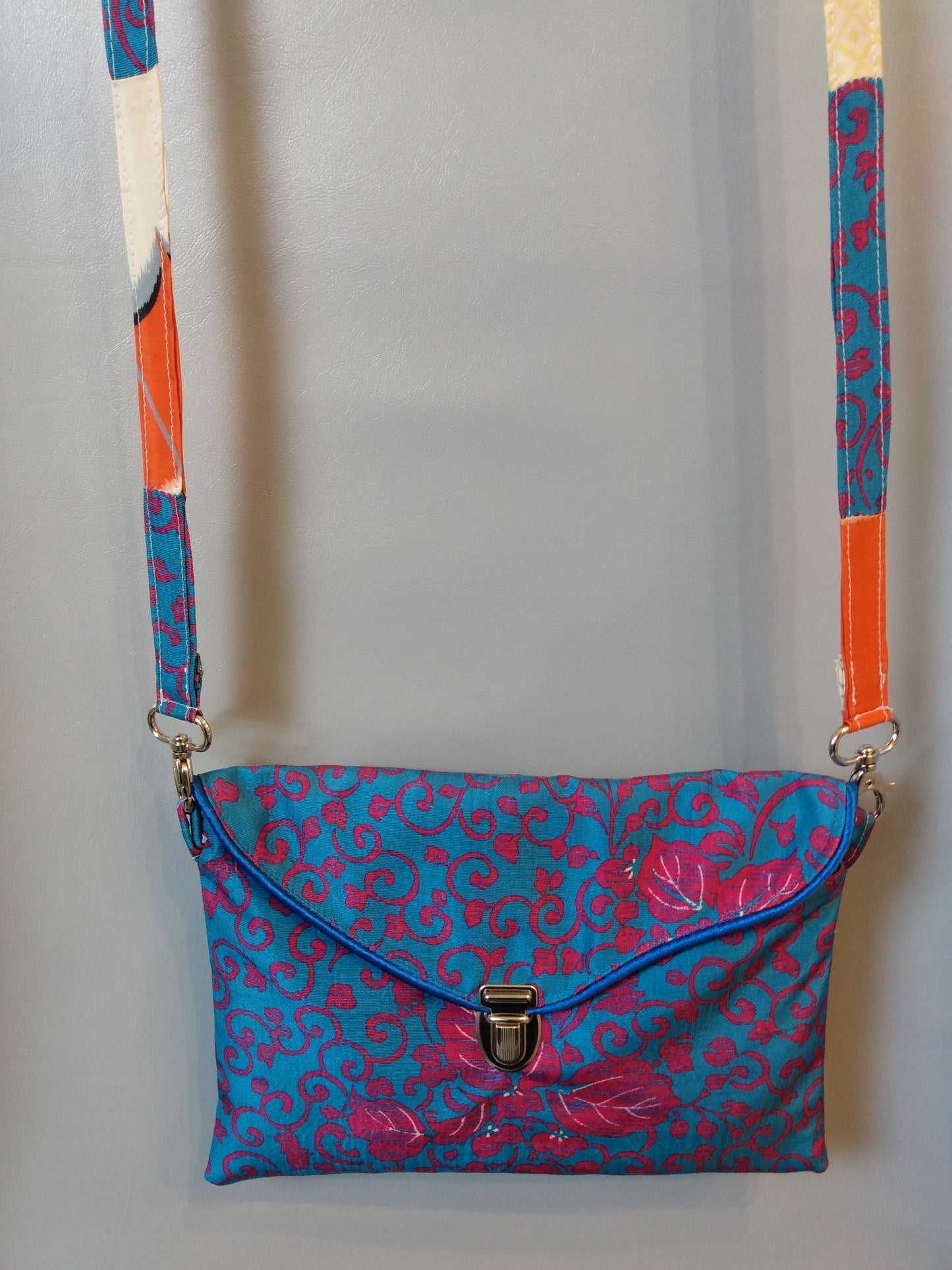 Avant de la pochette Kawa bleue et fuchsia. Pochette se fermant par un rabat et une attache cartable nickelée. Le rabat est passepoilé (bleu). Anse couleurs tissu extérieur et doublure.  tissu extérieur motifs floraux et arabesques fuchsia sur fond bleu clair. Doublure motifs grues blanches sur fond orange et beige.