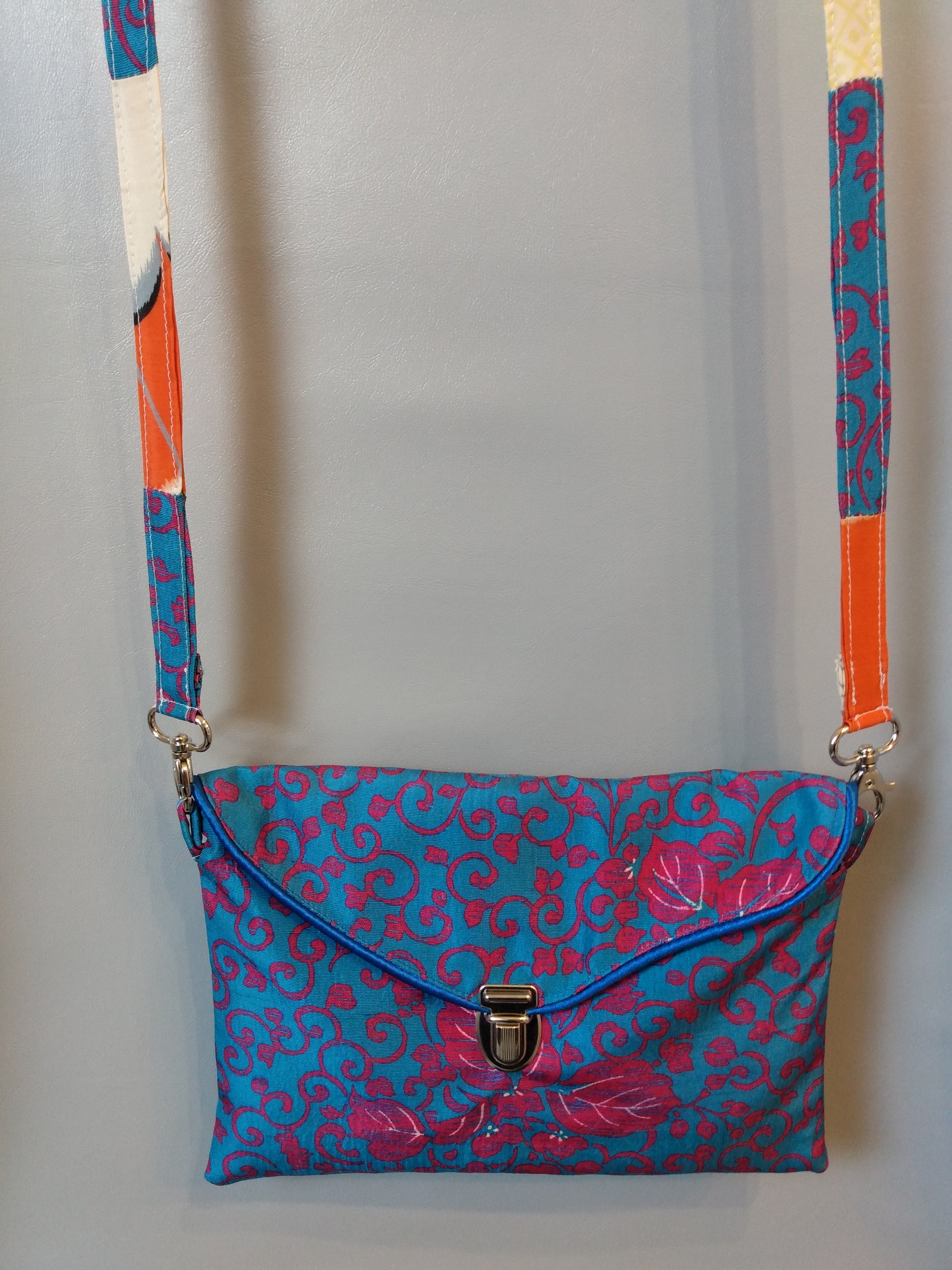 Avant de la pochette Kawa bleue et fuchsia. Pochette se fermant par un rabat et une attache cartable nickelée. Le rabat est passepoilé (bleu). Anse couleurs tissu extérieur et doublure.  tissu extérieur motifs floraux et arabesques fuchsia sur fond bleu clair. Doublure motifs grues blanches sur fond orange et beige.