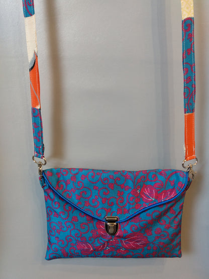 Avant de la pochette Kawa bleue et fuchsia. Pochette se fermant par un rabat et une attache cartable nickelée. Le rabat est passepoilé (bleu). Anse couleurs tissu extérieur et doublure.  tissu extérieur motifs floraux et arabesques fuchsia sur fond bleu clair. Doublure motifs grues blanches sur fond orange et beige.