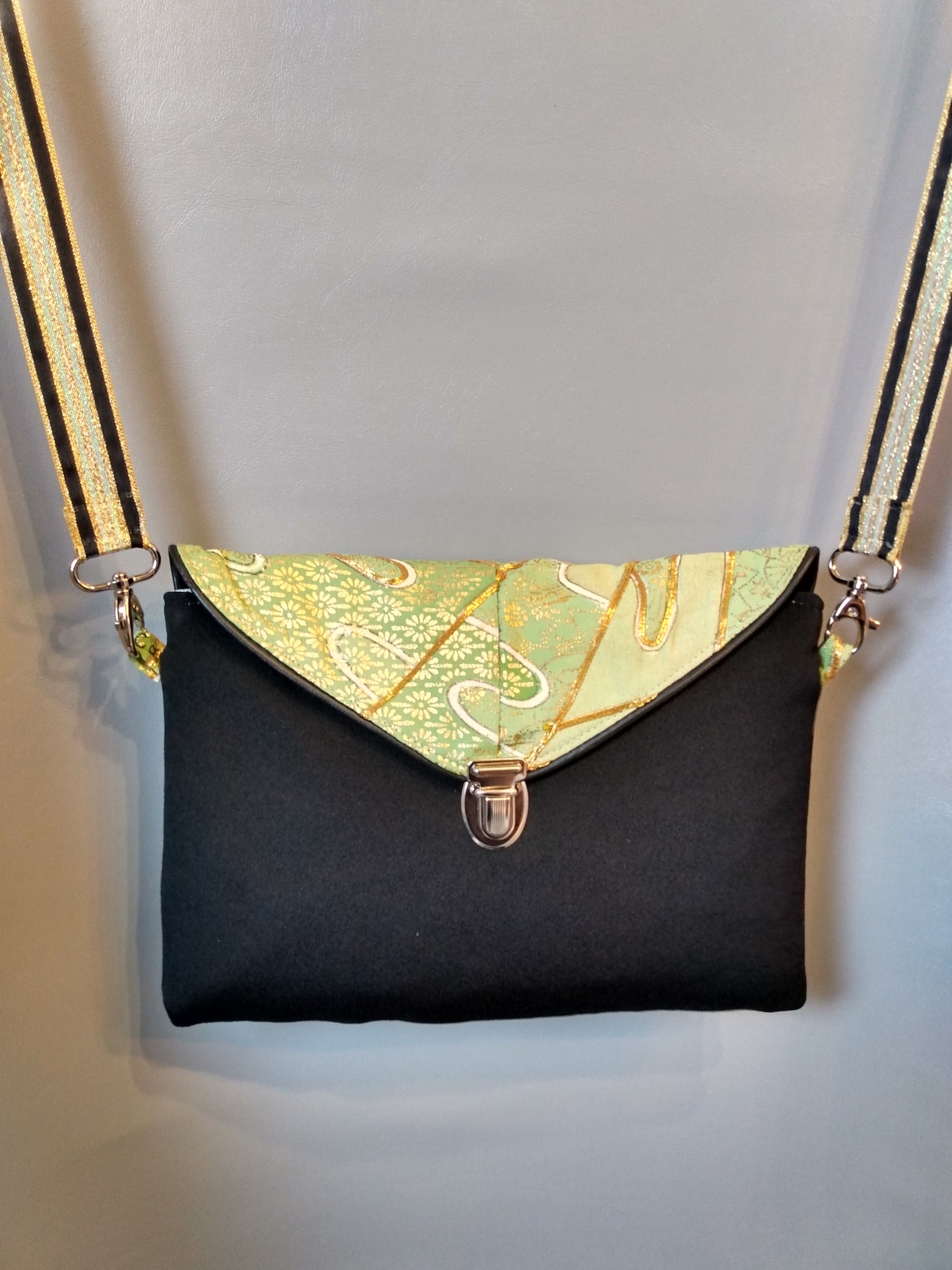Avant de la pochette Kawa vert et noir.
Pochette se fermant par un rabat et une attache cartable nickelée. Le rabat est de couleur verte et or et il est passepoilé (noir). Le corps du sac est noir. La bandoulière est noir, or et vert.