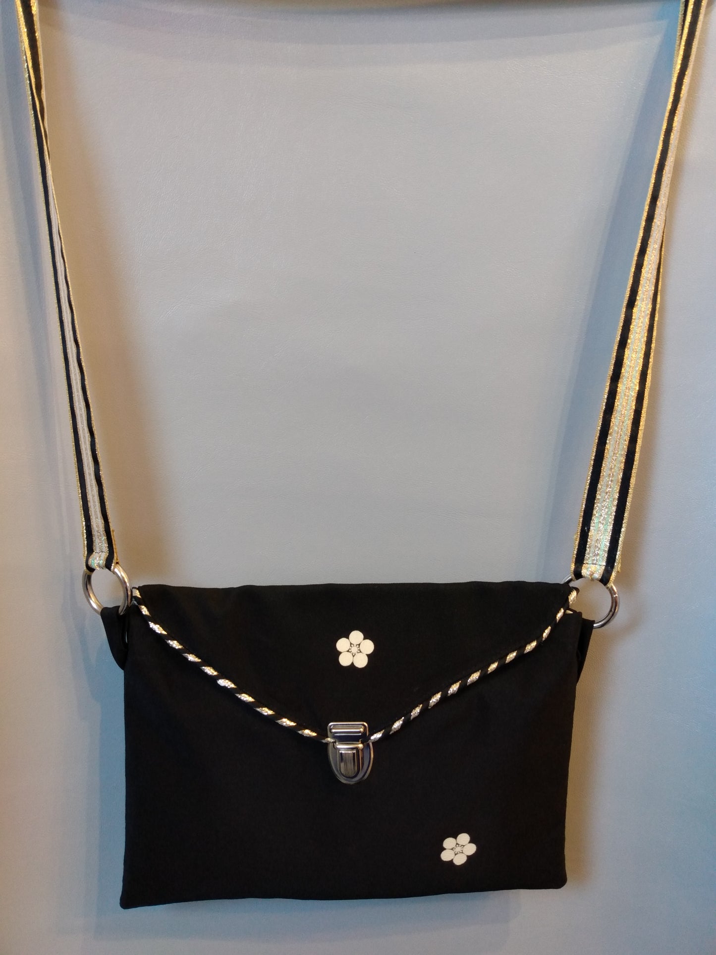 Avant de la pochette Kawa noir et fleur beige.
Pochette se fermant par un rabat noir avec une fleur beige au centre et une attache cartable nickelée. Le rabat est passepoilé (or et noir).
Le tissu extérieur est noir avec deux fleurs beiges.