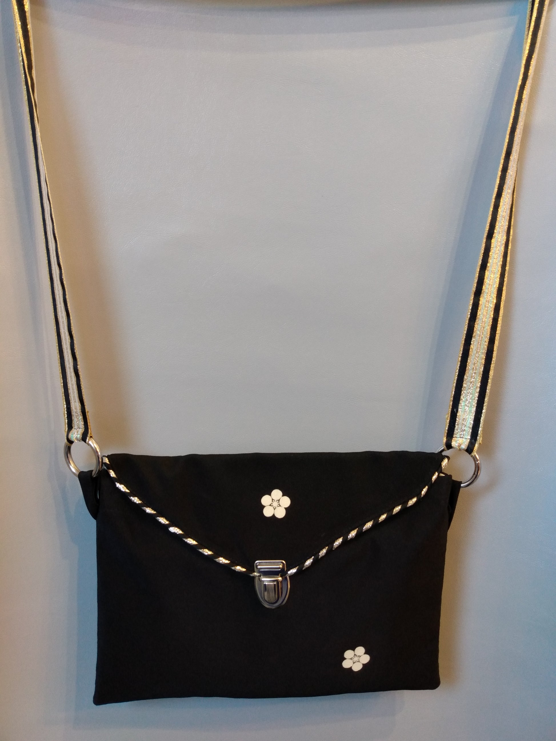 Avant de la pochette Kawa noir et fleur beige.
Pochette se fermant par un rabat noir avec une fleur beige au centre et une attache cartable nickelée. Le rabat est passepoilé (or et noir).
Le tissu extérieur est noir avec deux fleurs beiges.